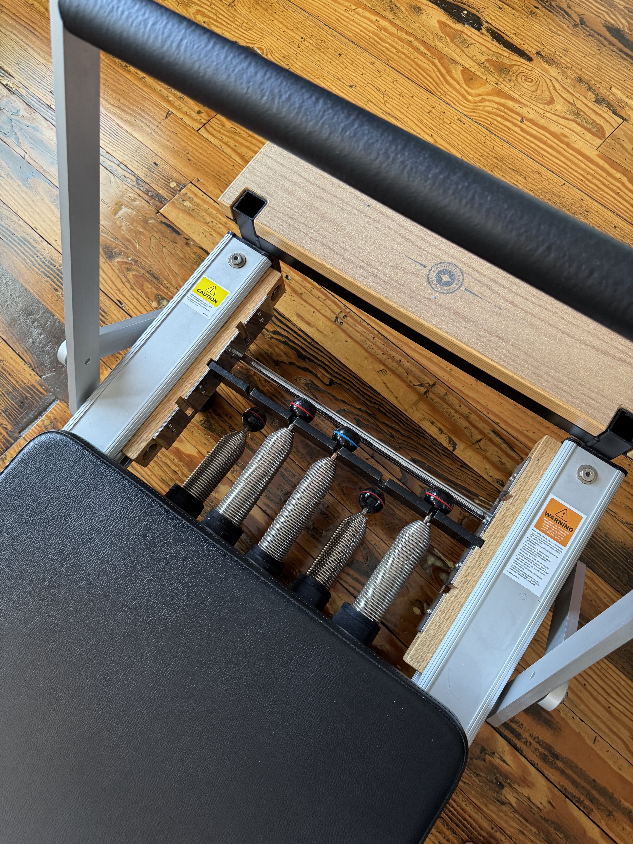 Pilates reformer with a black padded seat, a wooden platform, and a metal frame with springs and adjustable supports, placed on a wooden floor.