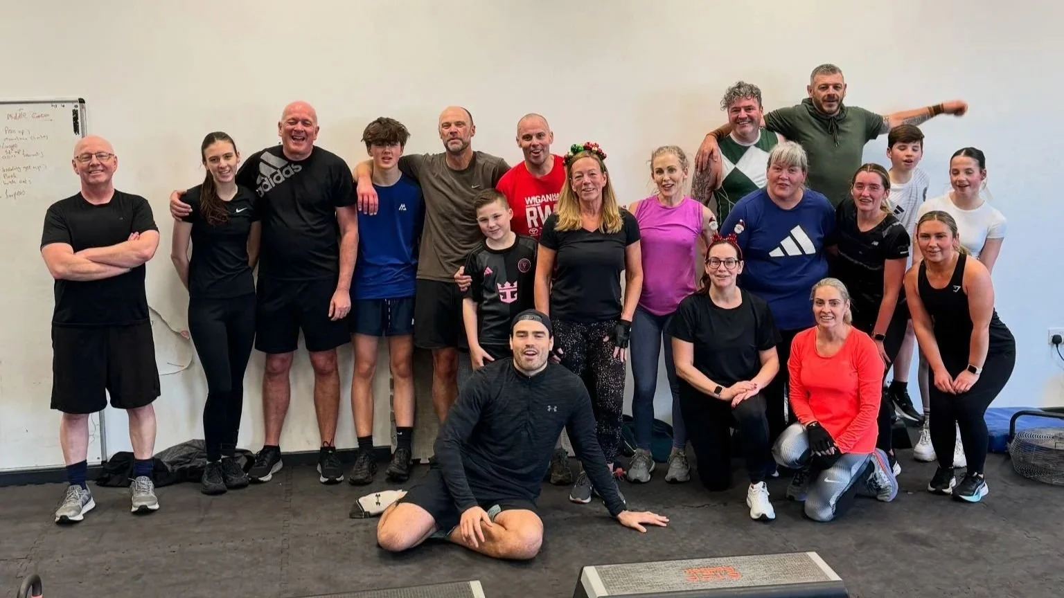 Group of people in workout attire posing together indoors, some smiling and others making playful poses, on a carpeted floor with a whiteboard on the wall.