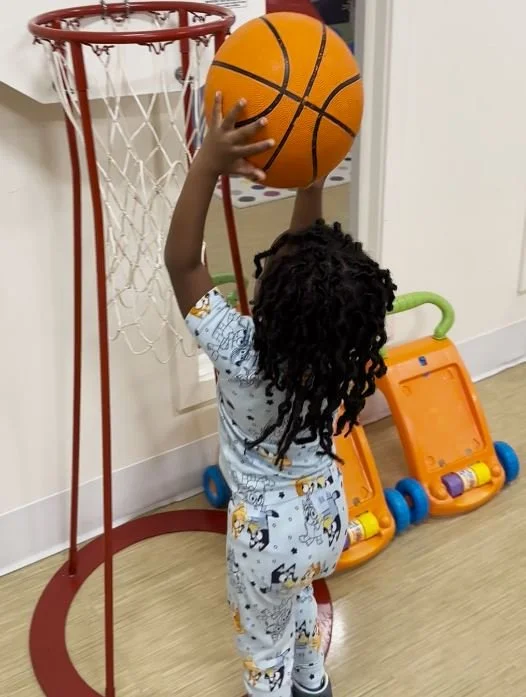 Child reaching up to dunk a basketball into a miniature basketball hoop in a playroom.