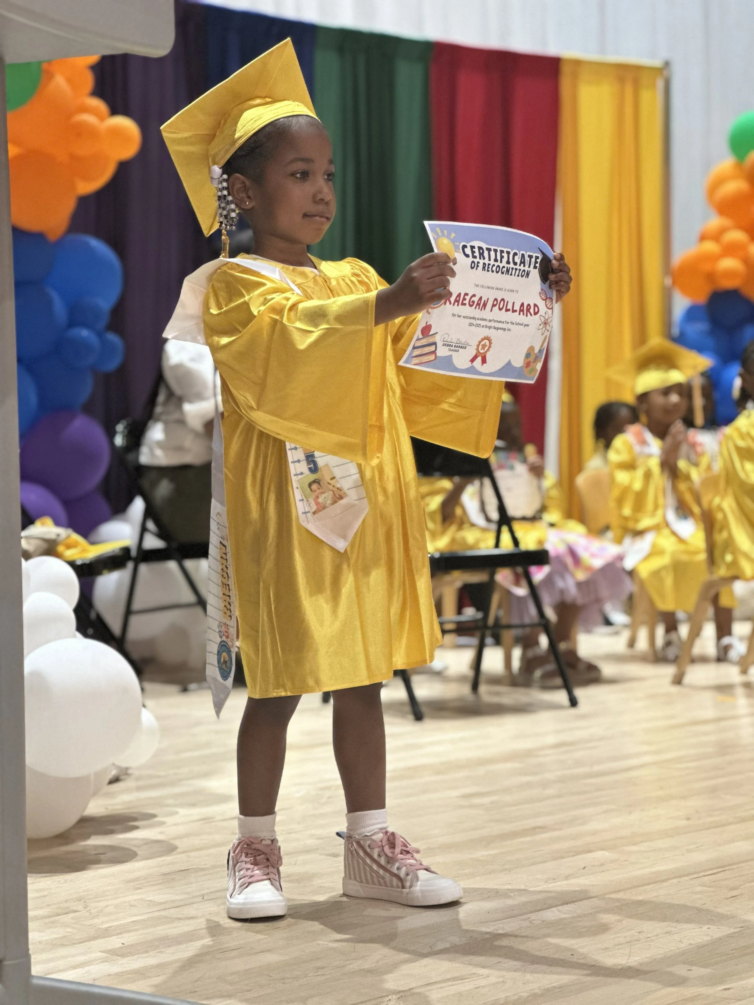 A young girl in a yellow graduation gown and cap holding a certificate on stage during a graduation ceremony. She stands on a wooden floor with colorful balloon decorations and other children dressed in similar caps and gowns in the background.