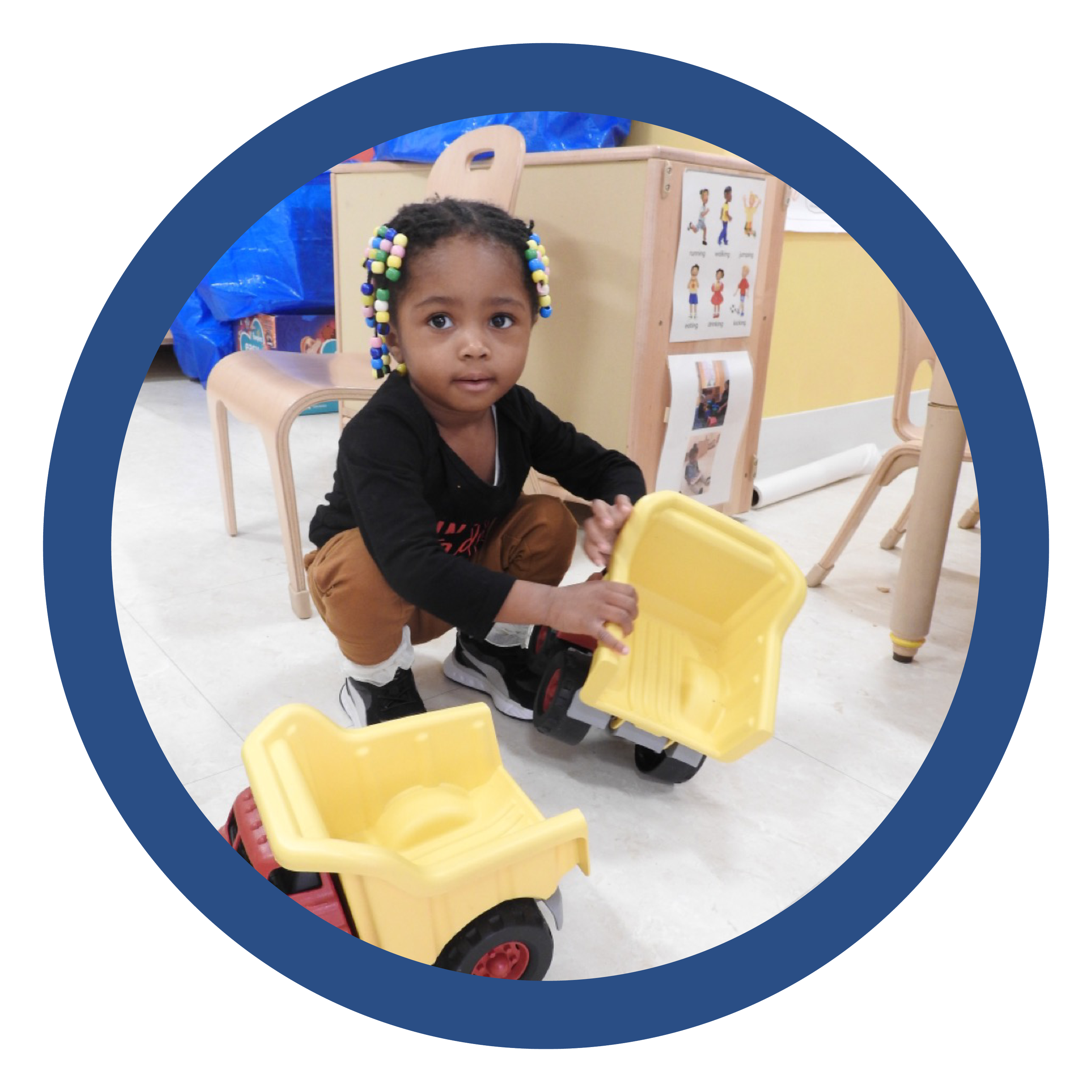 A young girl with colorful beaded hair playing with yellow toy dump trucks in a classroom.
