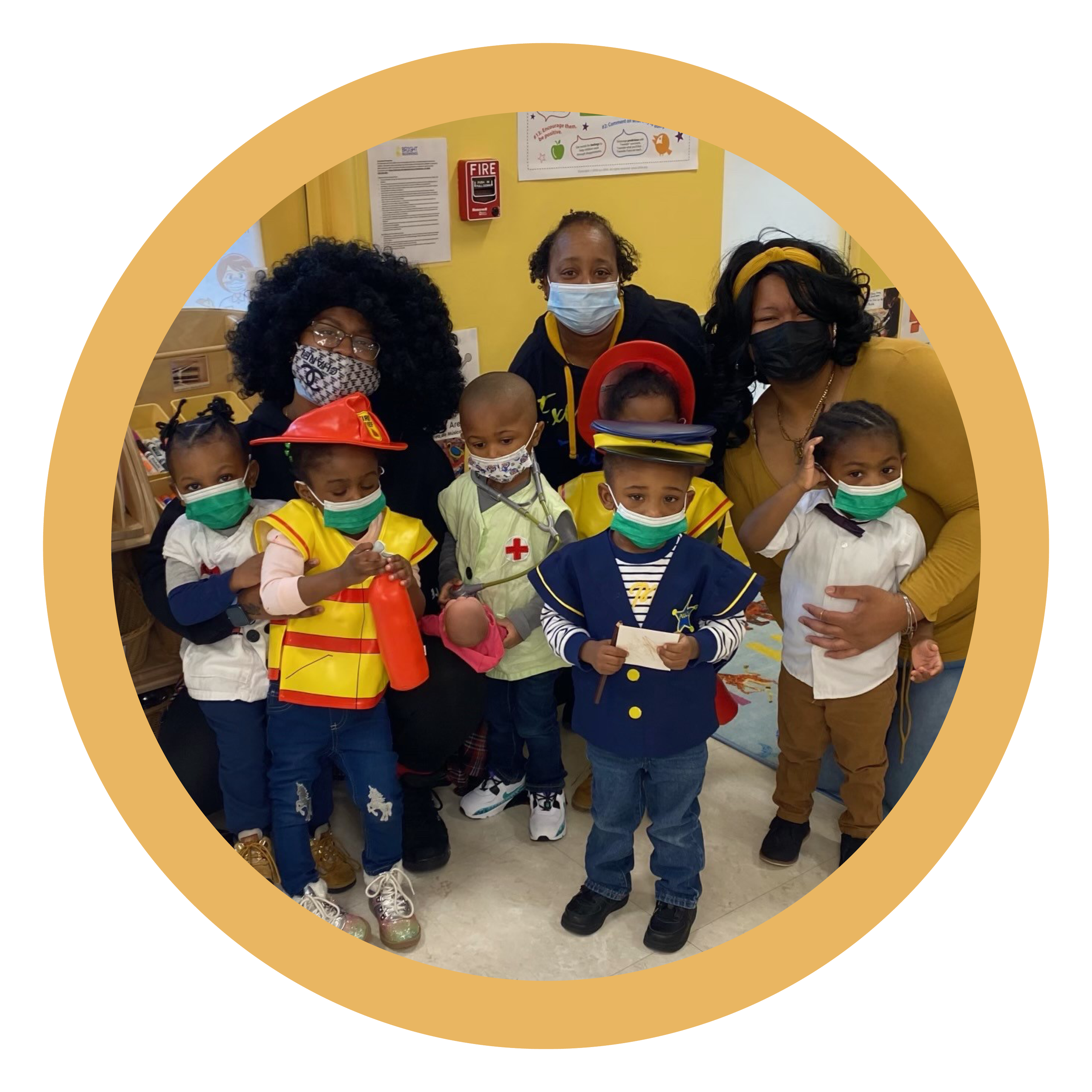 Group of young children in costumes and face masks with three adult women in masks, inside a classroom.