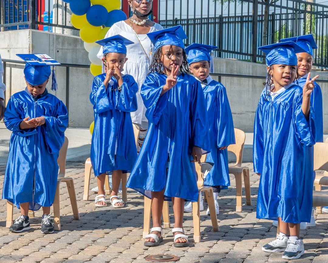 A group of young children dressed in blue graduation gowns and caps, standing outdoors with some sitting on small chairs, with a woman in white and balloons in the background.