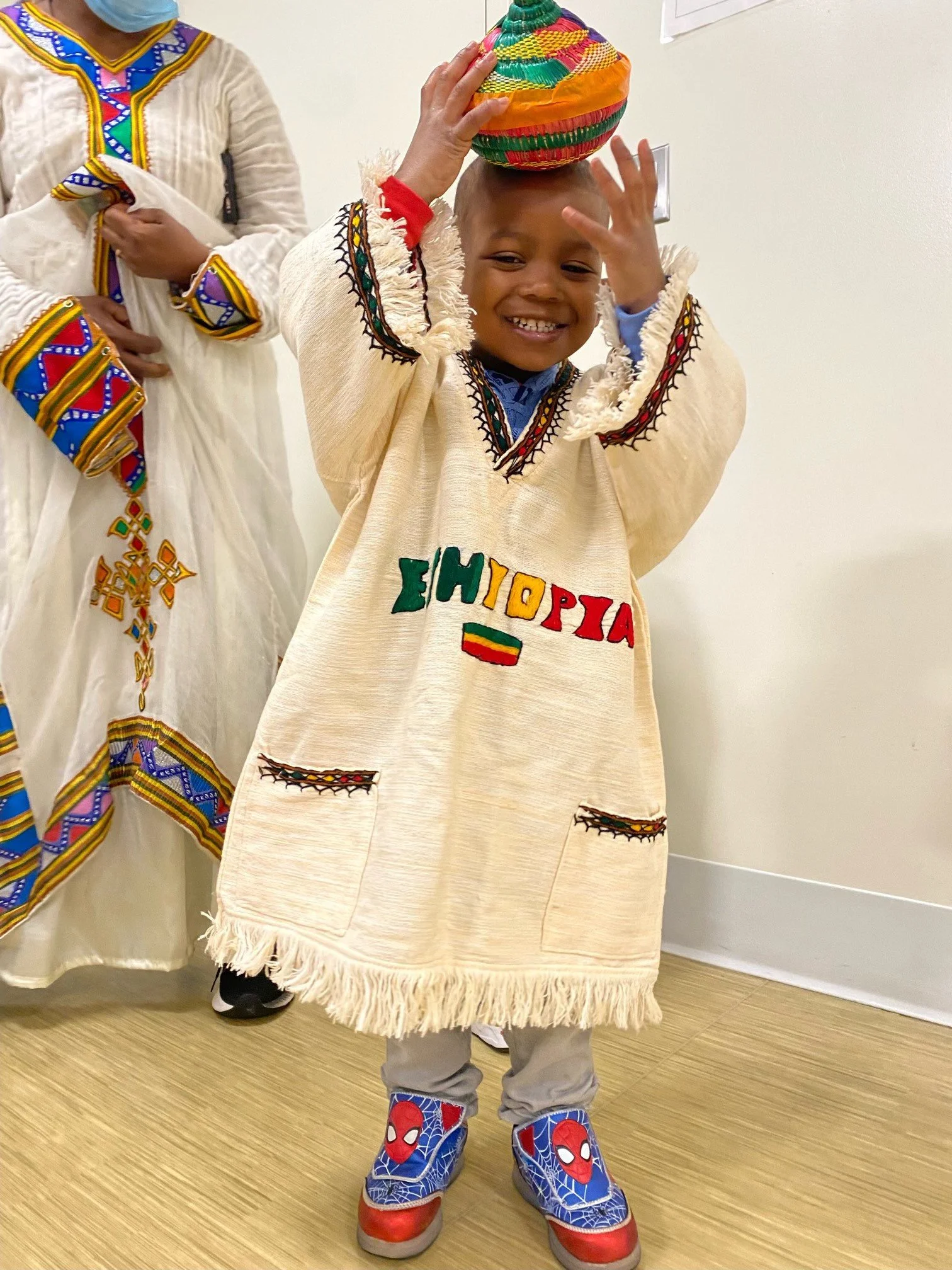 A young boy smiling and playing with a colorful woven basket on his head, wearing a traditional Ethiopian garment with the word Ethiopia written on it, and Spider-Man shoes, standing against a white wall.