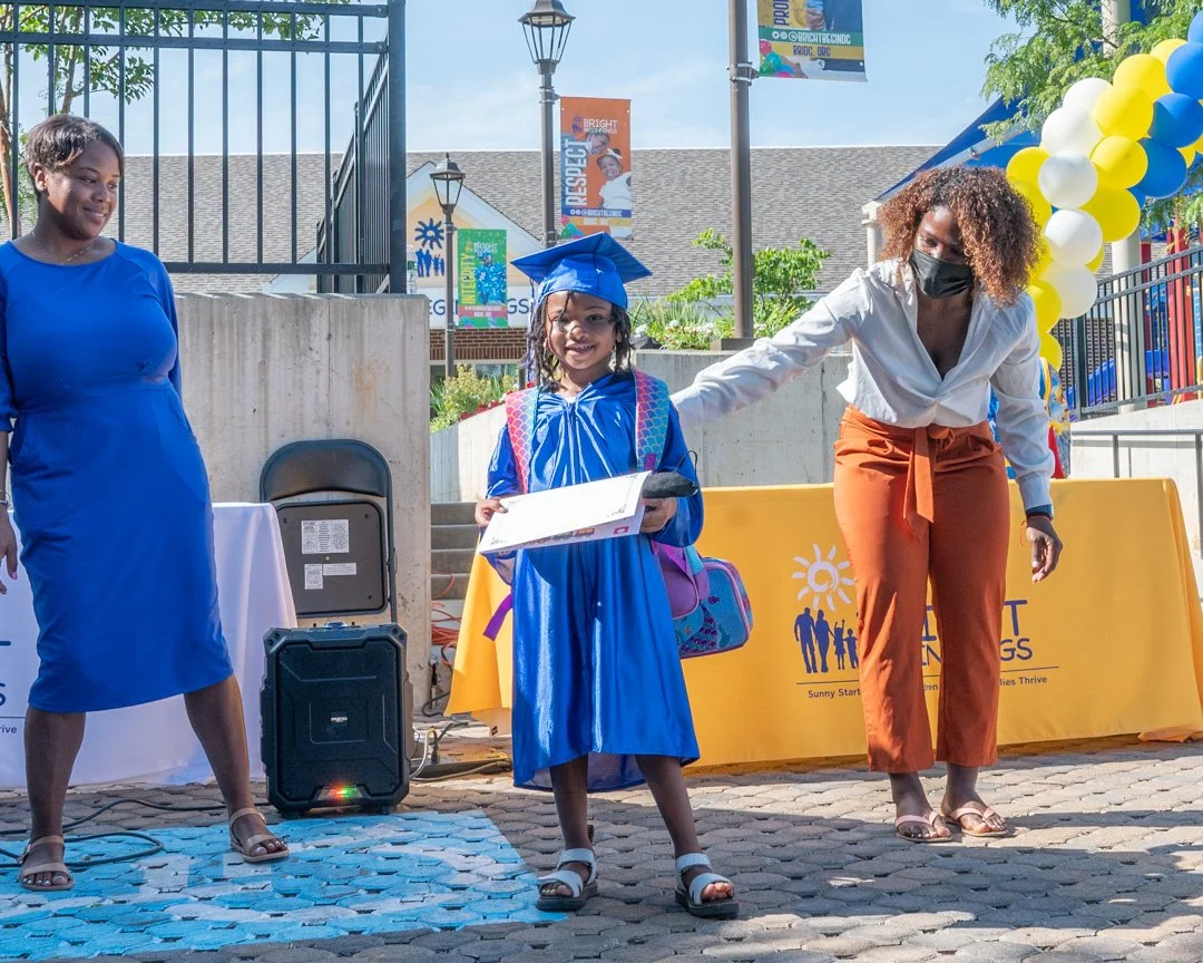 Young girl in a blue graduation cap and gown holding a diploma, smiling while standing on cobblestone at outdoor event with woman in white blouse and orange pants, and another woman in blue dress nearby, with yellow and blue balloons and a table in t