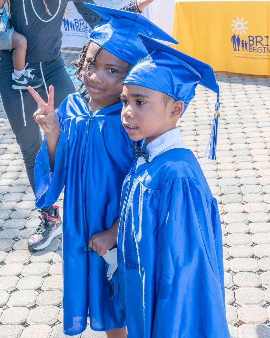 Two children in blue graduation caps and gowns at their graduation ceremony, one making a peace sign and the other looking forward.