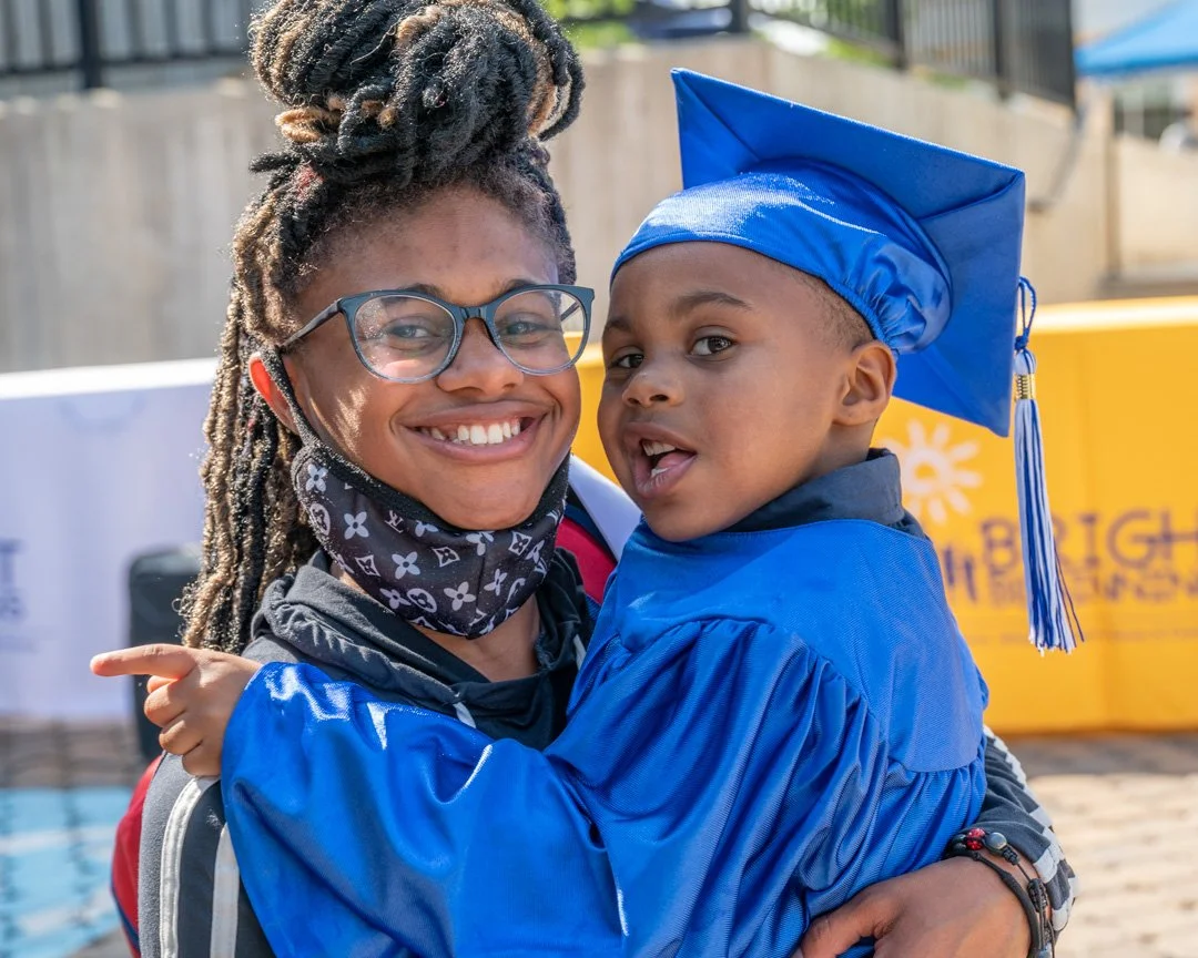 A woman with glasses and dreadlocks holding a young boy in a blue graduation gown and cap.