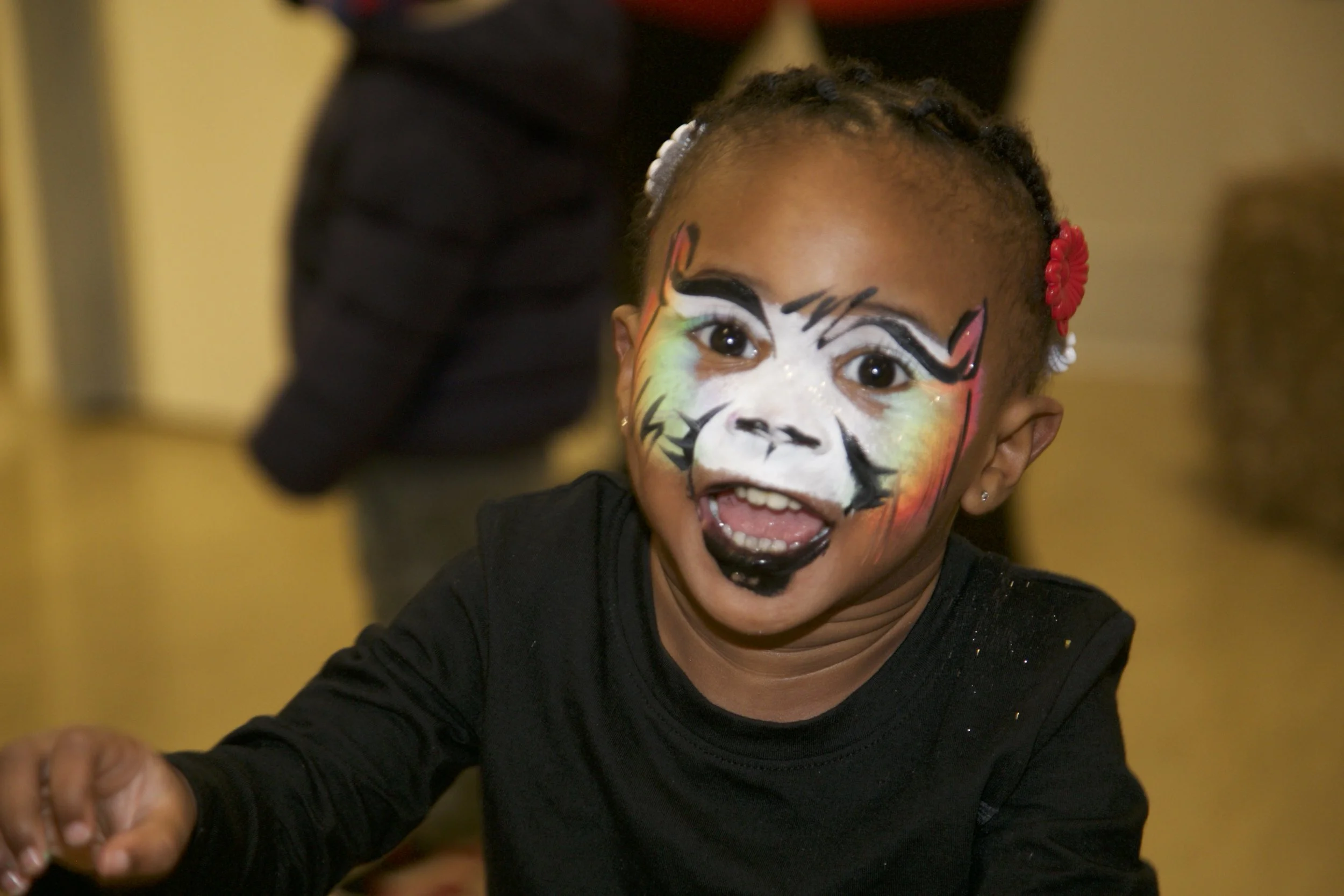 A young girl with face paint resembling a colorful tiger, smiling with her mouth open and showing her teeth.