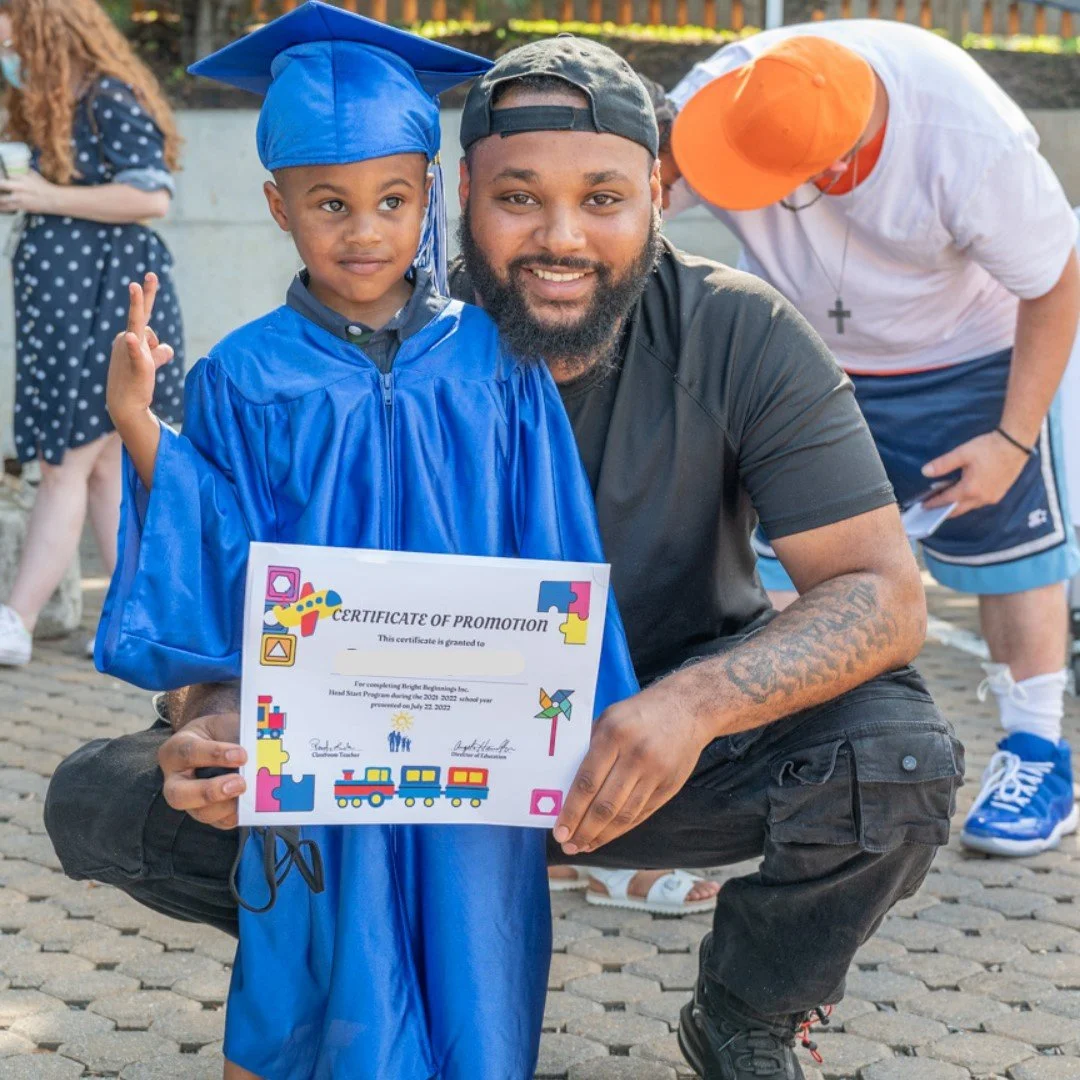 A young boy in a blue graduation gown and cap holding a certificate, posing with a man kneeling beside him during a graduation celebration outdoors.