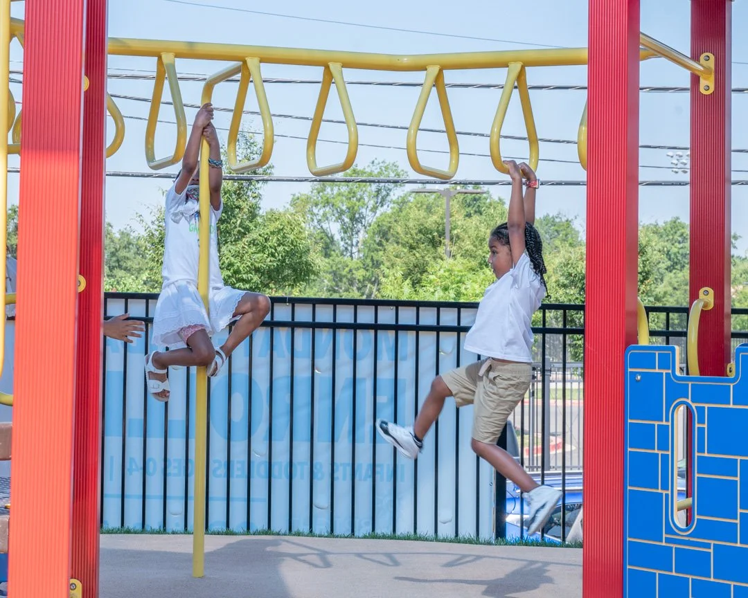 Two children hanging from playground bars at a playground with trees and a fence in the background.