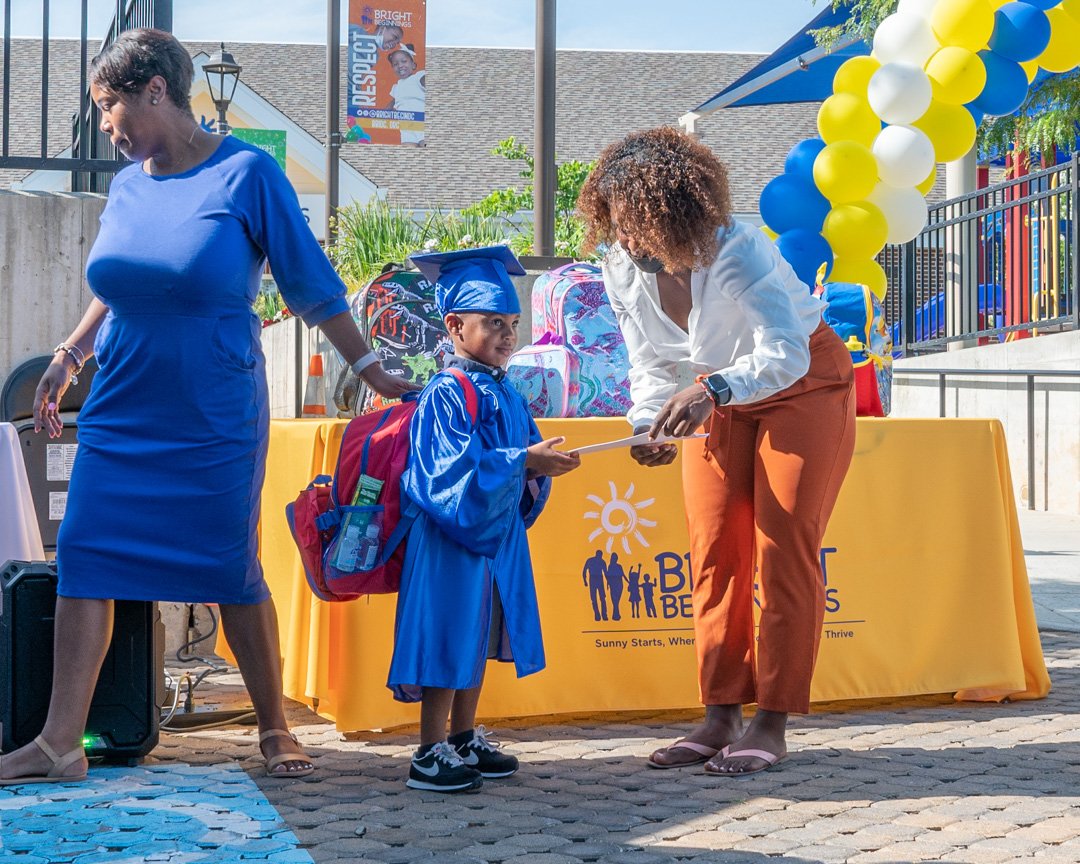 A young boy in a blue graduation cap and gown receiving a diploma from a woman at an outdoor graduation event, with another woman nearby and yellow, blue, and white balloons in the background.