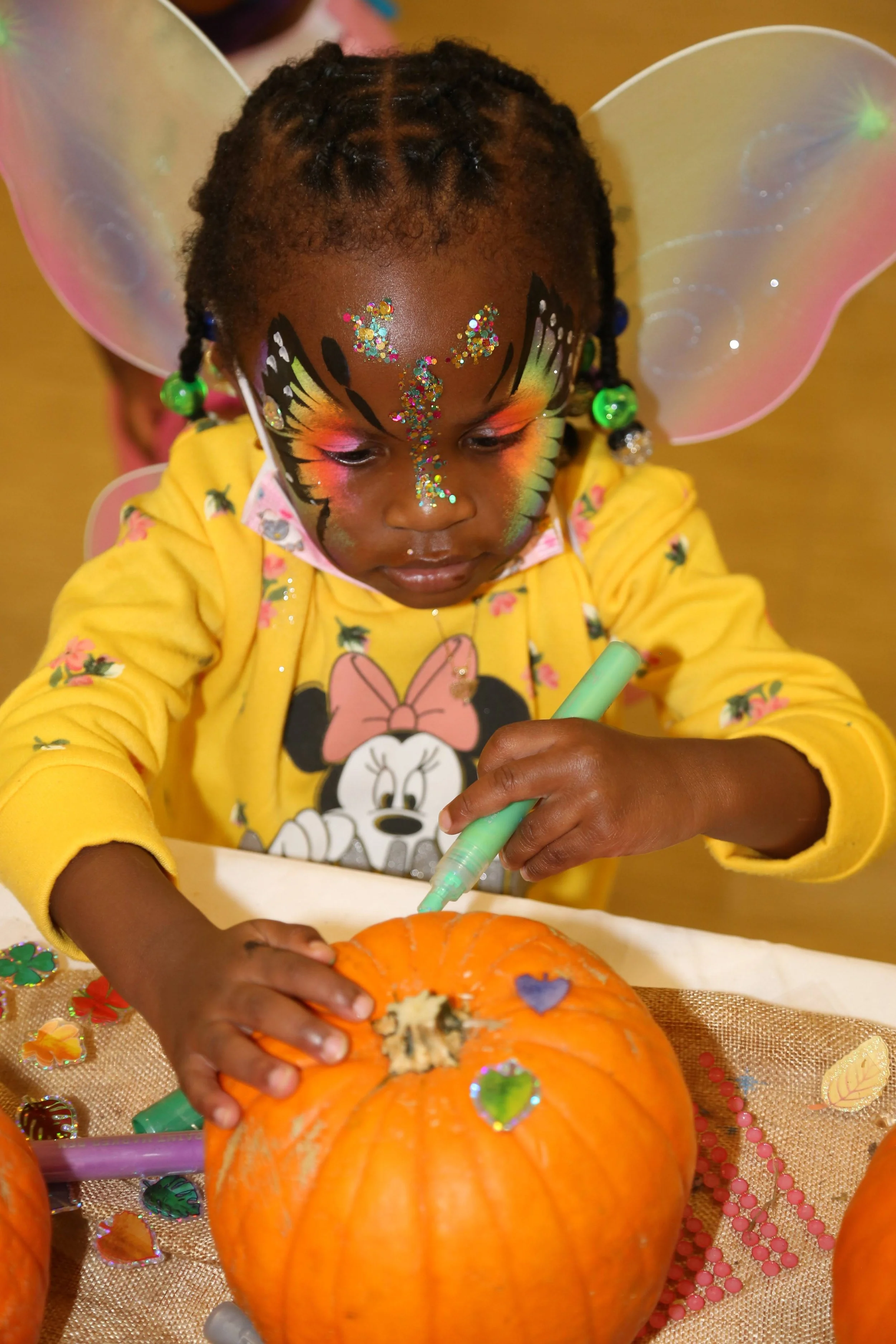 A young girl with face paint and fairy wings decorating a pumpkin at a craft table.