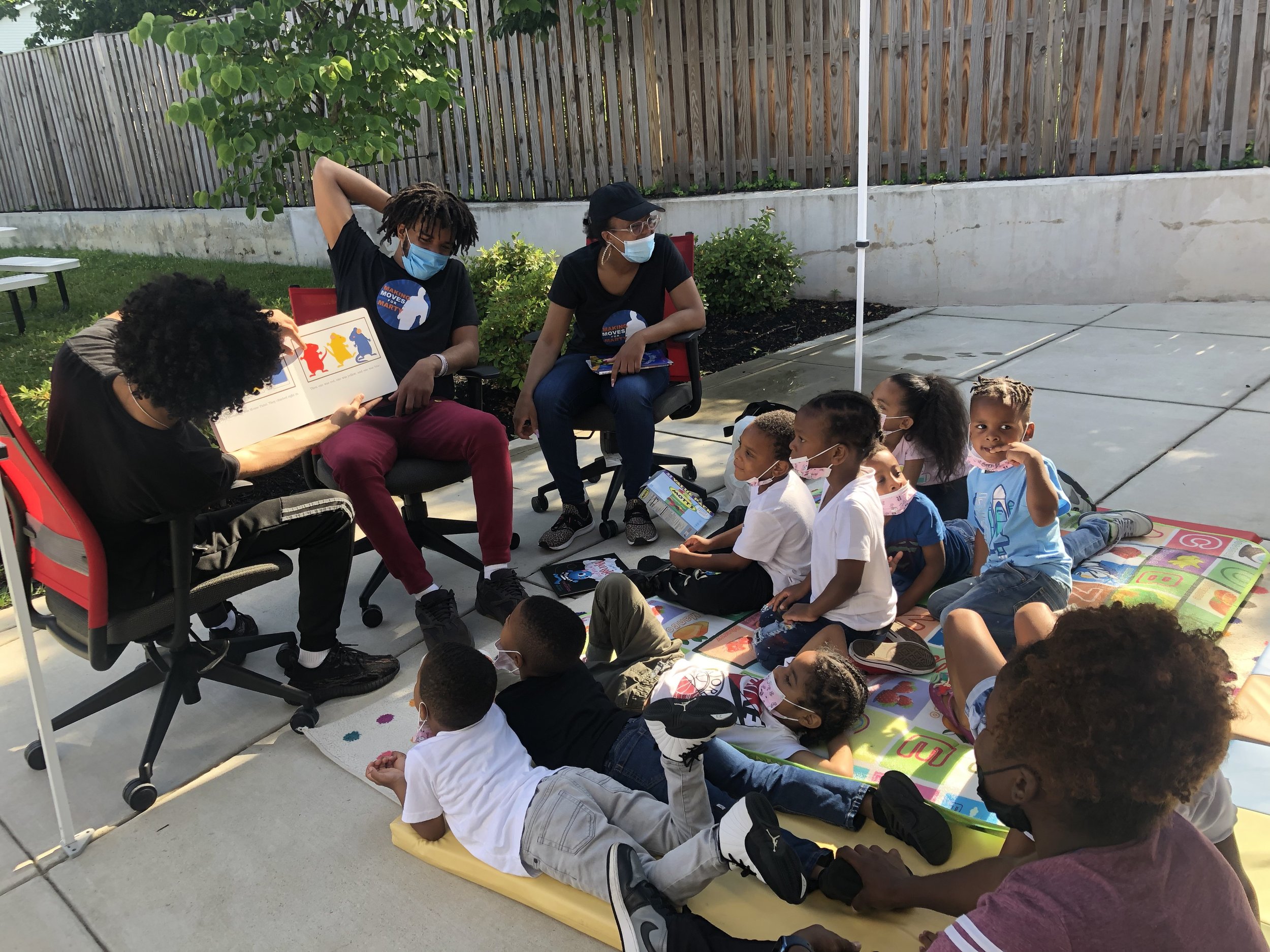 Group of children and adults sitting outdoors on a blanket and chairs, listening to a person reading a children's book. The children are wearing masks, and the background includes a wooden fence, greenery, and a white pole.