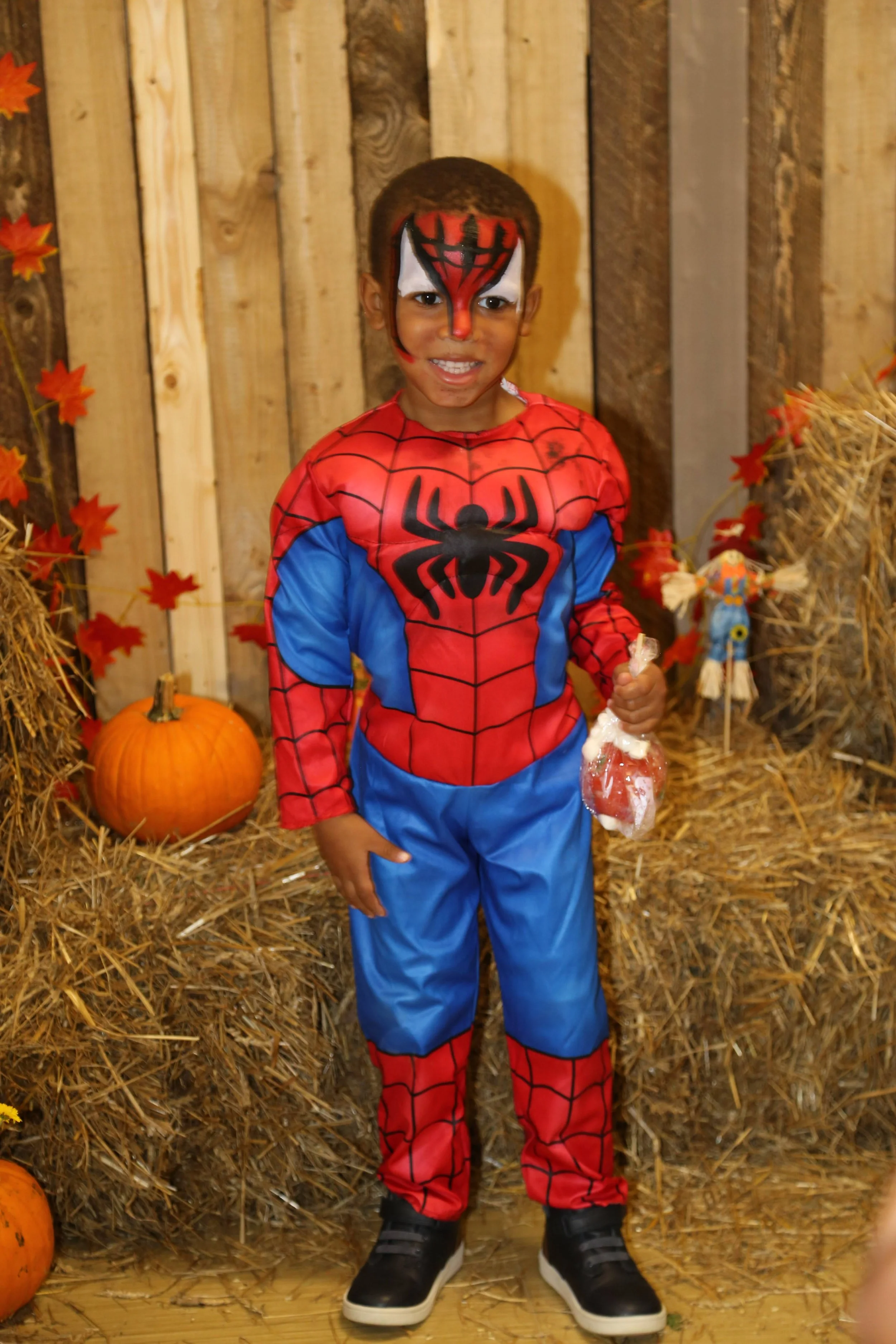 Young boy dressed as Spider-Man holding a treat, standing in front of hay bales, pumpkins, and fall decorations.