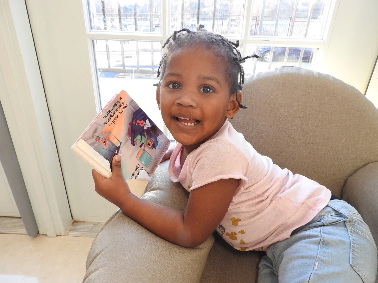 A young girl with braided hair and a pink t-shirt, lying on a beige couch, smiling and holding a children's book. Behind her is a large window with sunlight streaming in.