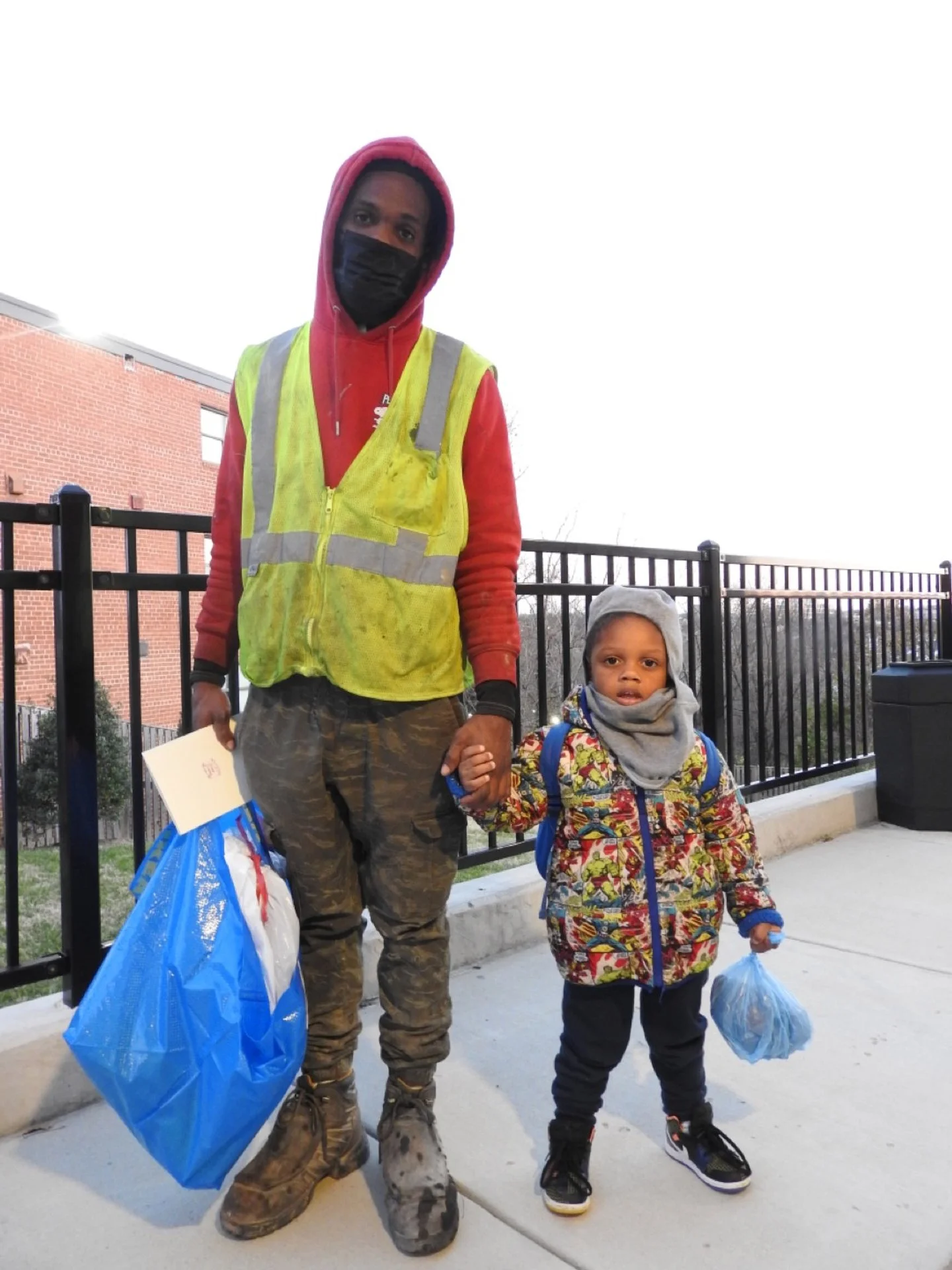 A man and a young boy standing outdoors on a sidewalk, holding hands. The man is wearing a red hoodie, a yellow reflective safety vest, camouflage pants, boots, and a black face mask. The boy is wearing a colorful jacket, gray hat, and gray hoodie, h