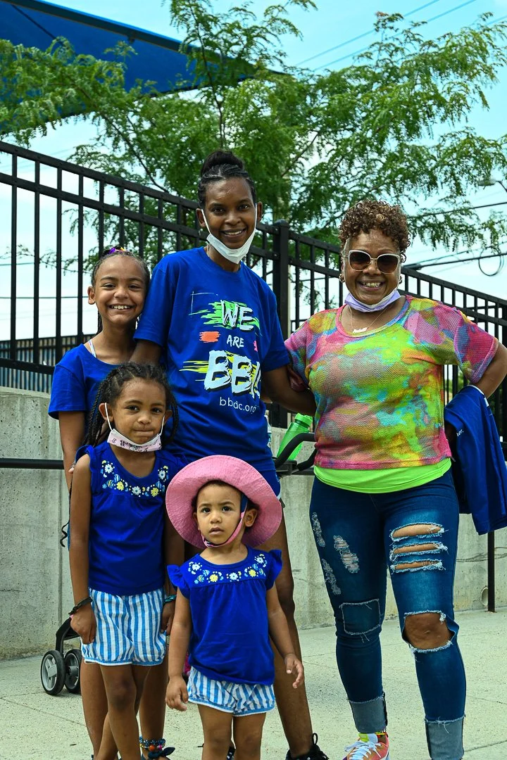 A group of five smiling females, including three young girls and two adult women, standing outdoors in front of a black fence with trees in the background. They are wearing colorful clothing, with some wearing masks pulled down below their chins. The