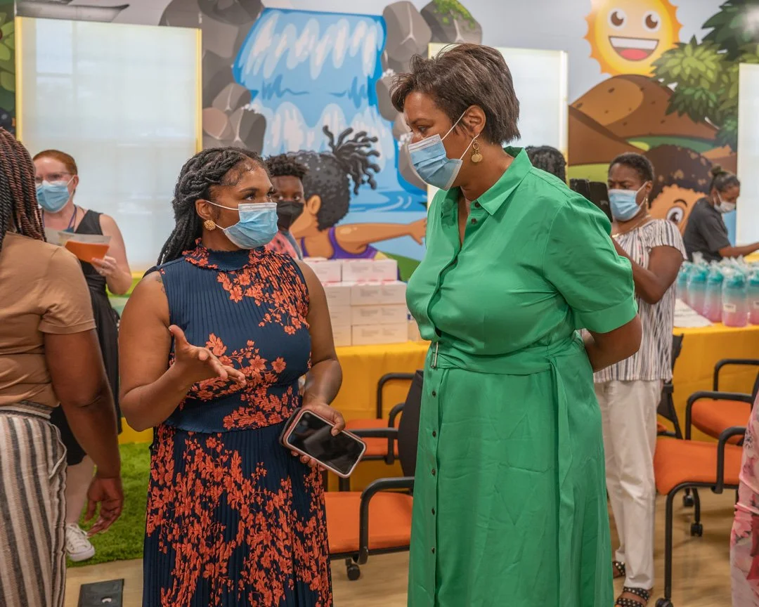 Two women wearing face masks are engaged in a conversation in a colorful indoor setting with other people in the background. One woman is wearing a navy blue dress with an orange floral pattern, and the other is in a bright green dress.