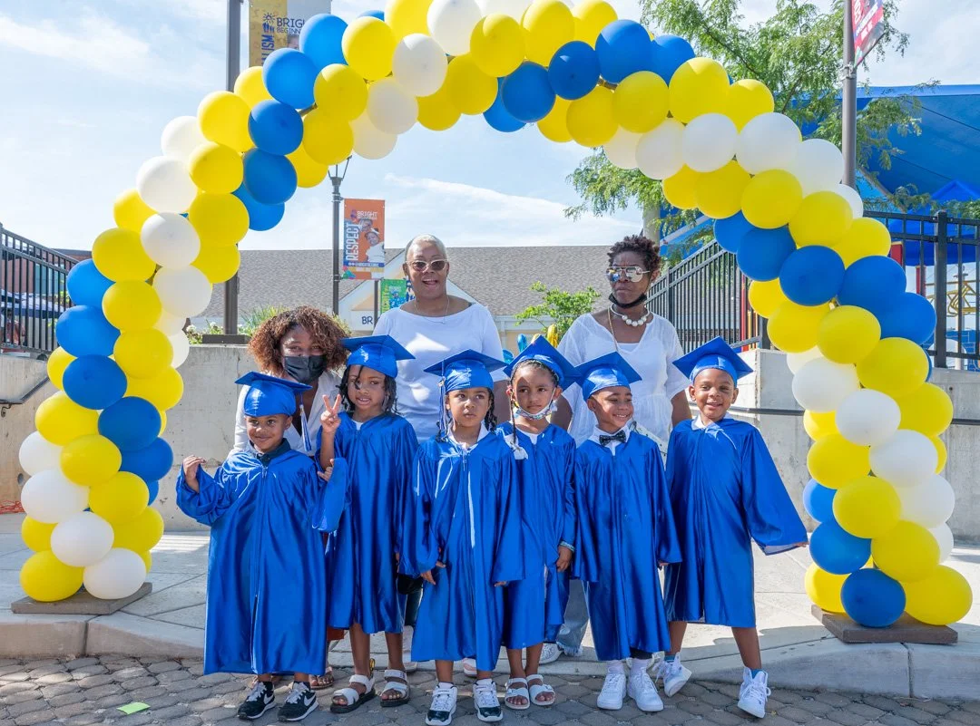 Group of young children in blue graduation gowns and caps standing under a balloon arch with three women behind them, outdoors on a sunny day.