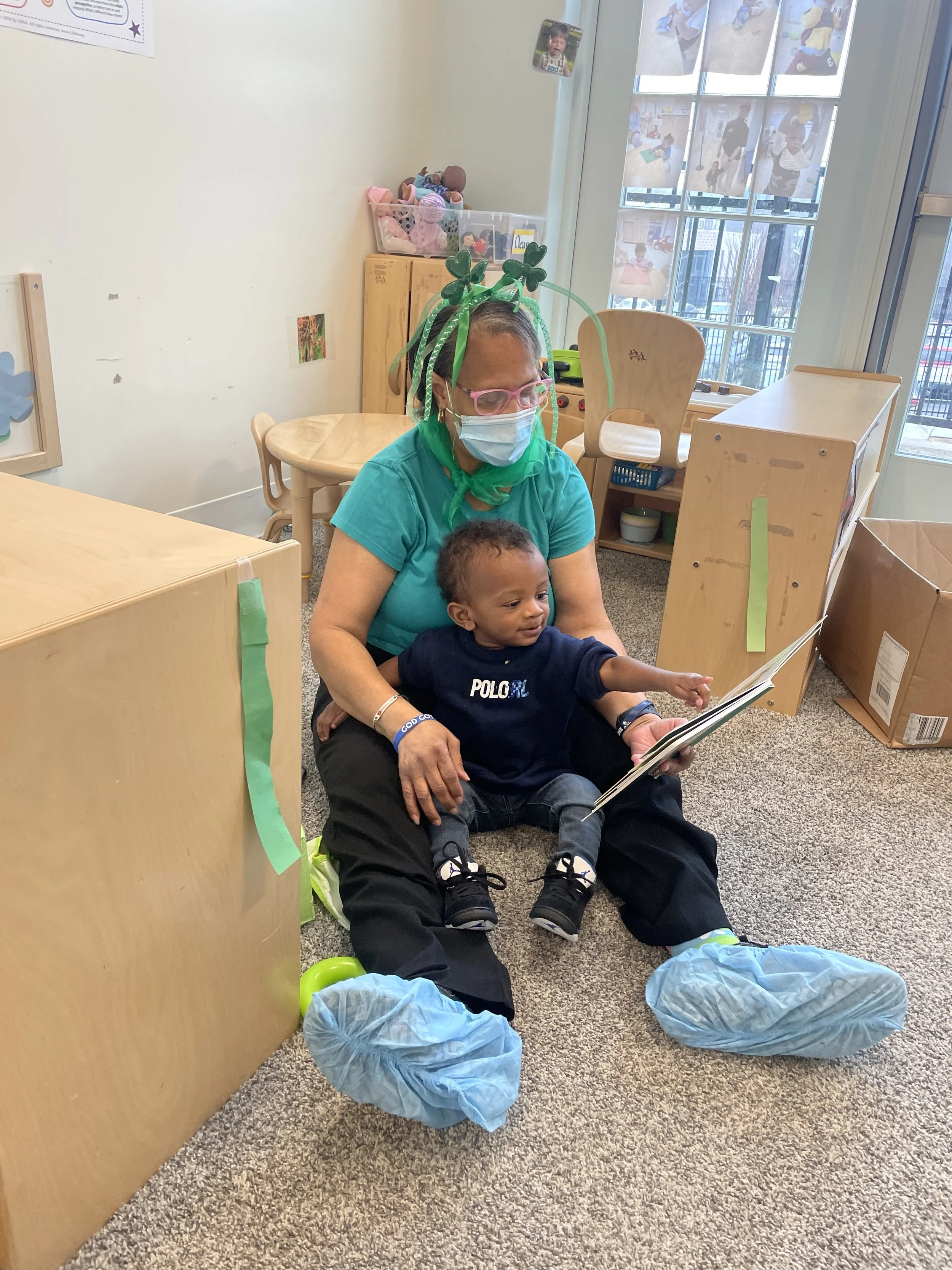 A woman wearing a face mask, glasses, and green-themed headband, sits on the floor reading a book to a young boy in a blue Polo shirt and jeans in a classroom setting.