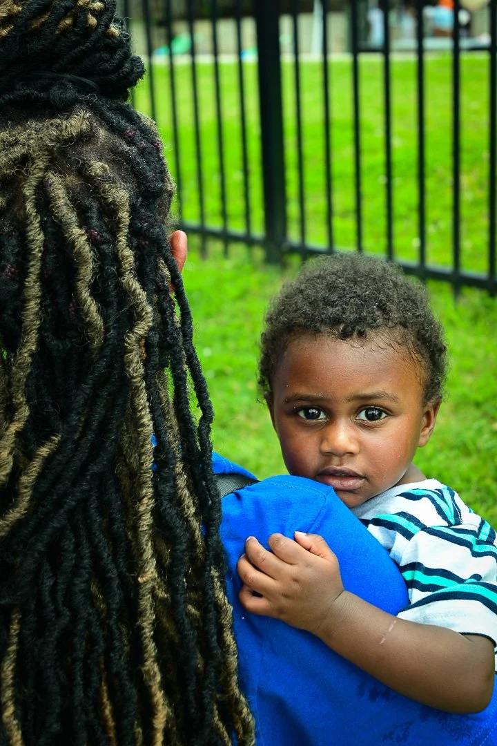 A young child with curly hair being held by an adult with dreadlocks outside near a black metal fence and green grass.