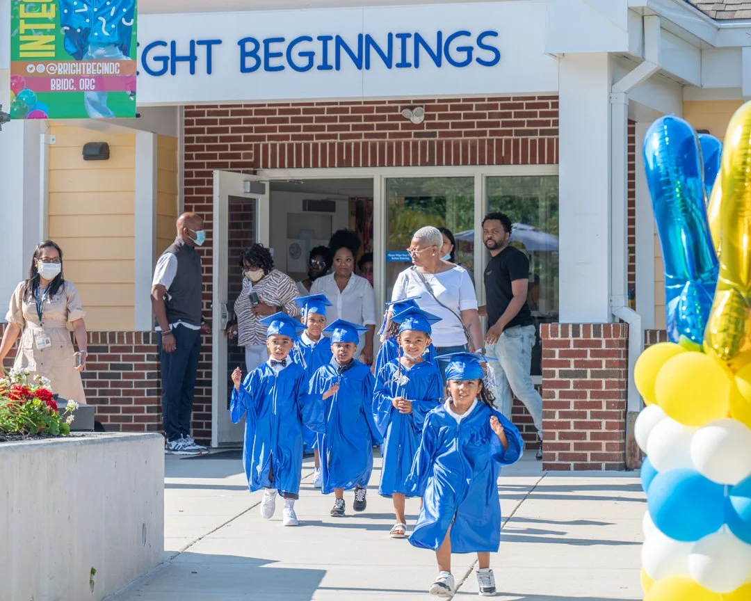 A group of young children in blue graduation caps and gowns walking out of a building labeled 'Night Beginnings'. They are accompanied by adults, some wearing masks, celebrating a graduation event.