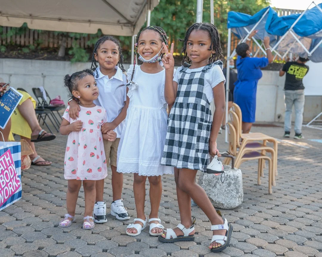 Four young girls standing together outdoors, smiling and making peace signs, with two women in the background, one adjusting a large blue tent. The girls are wearing casual dresses and sandals, and one has a face mask around her neck. The setting app