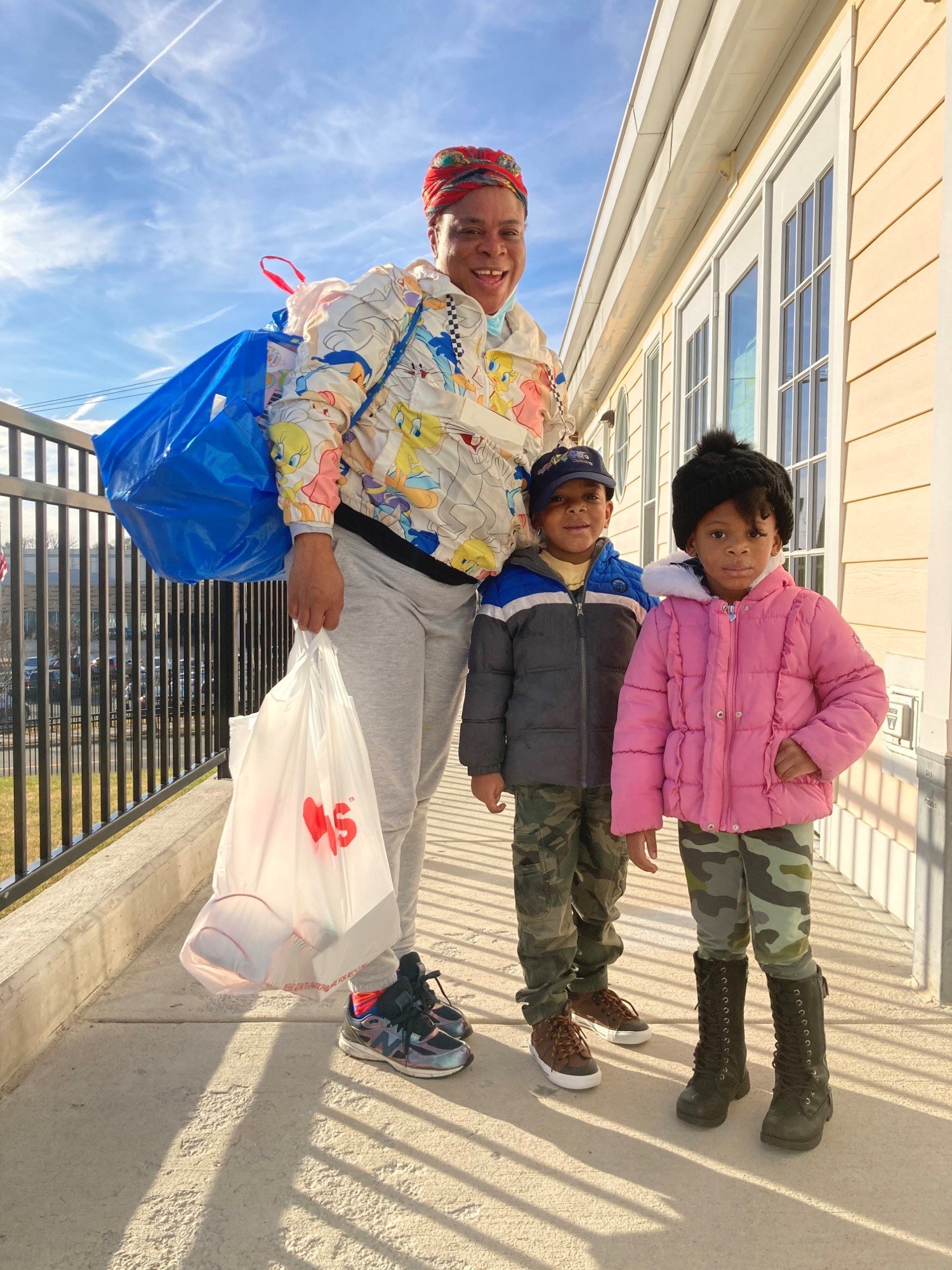 A woman and two children standing outside on a sidewalk, smiling at the camera. The woman is wearing a colorful jacket with cartoon characters, a red headscarf, and holding a shopping bag. The children are dressed in warm jackets, one in pink and cam