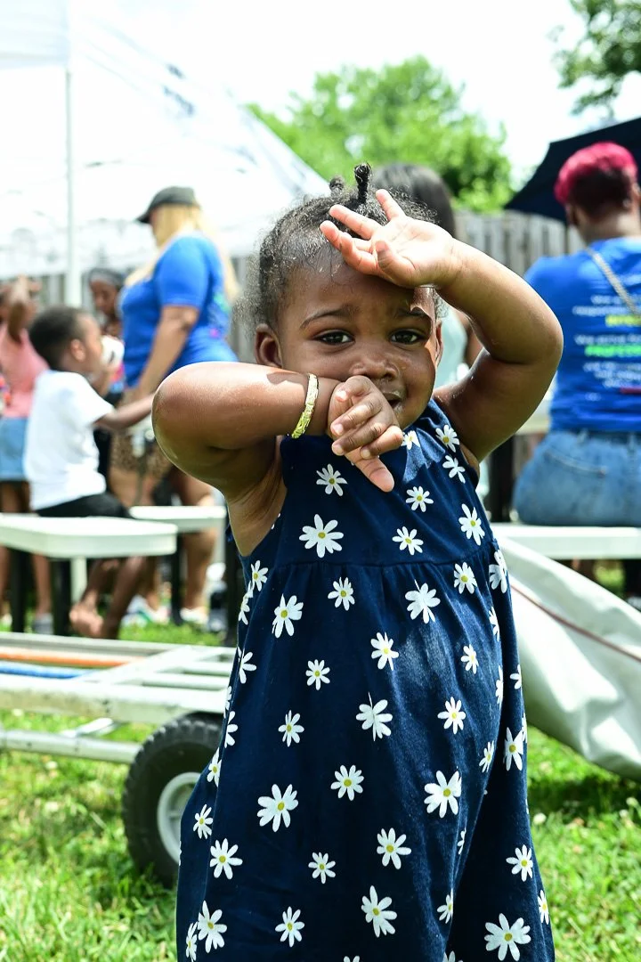 A young girl with dark skin and braided hair, wearing a navy blue dress with white daisy prints, is outdoors at a gathering, making a playful gesture with her hand near her forehead and face, with tents, people, and trees in the background.