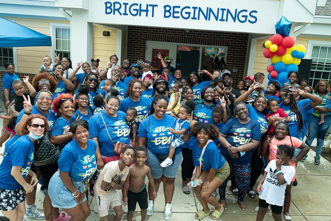A large group of smiling people, including children and adults, gathered in front of a building with a sign that says 'Bright Beginnings.' They are celebrating, with some making peace signs, and many wearing matching blue shirts with colorful text an