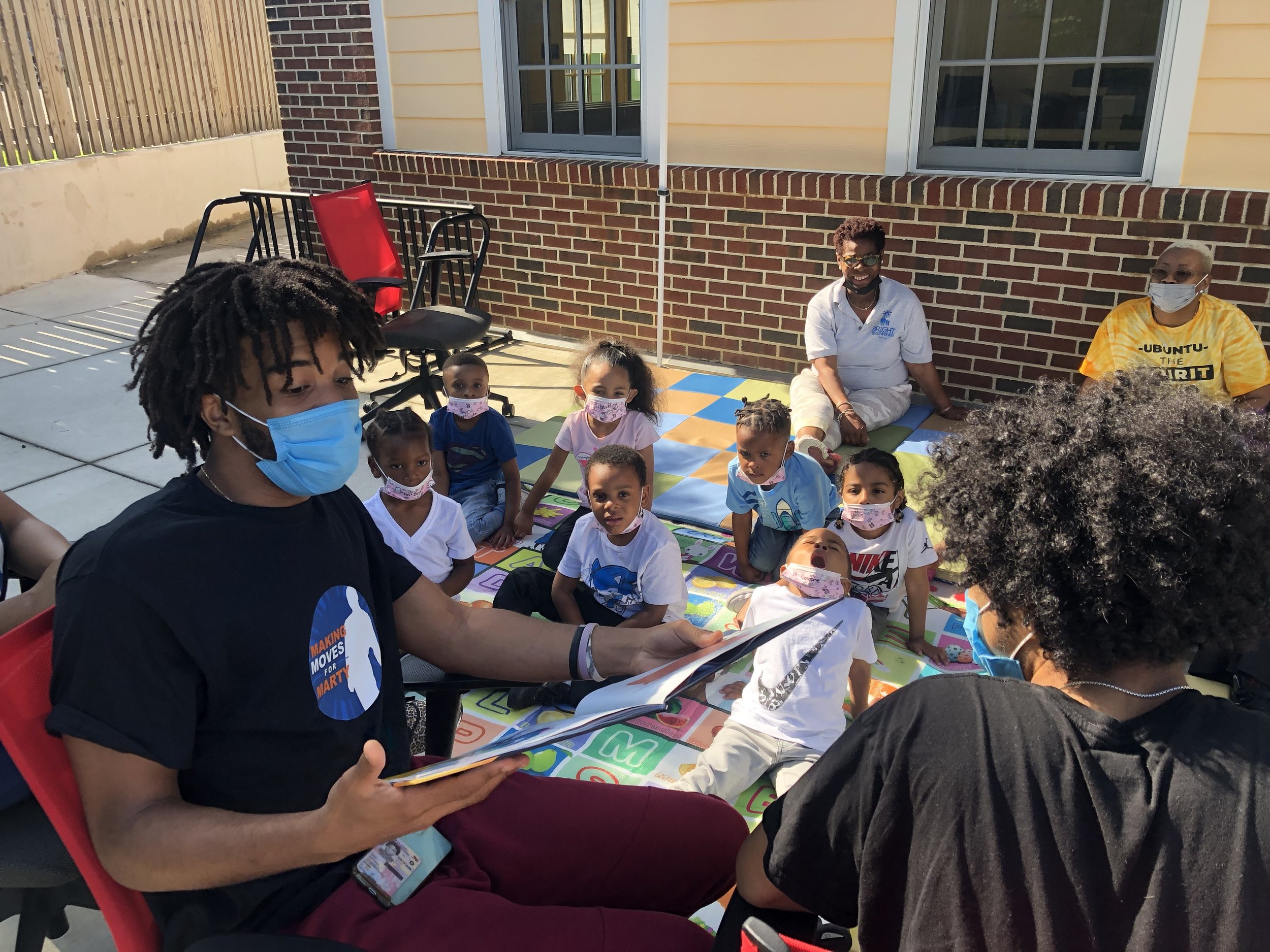 Children and adults sitting outdoors on colorful mats, wearing face masks, participating in a group activity near a brick wall and yellow house.