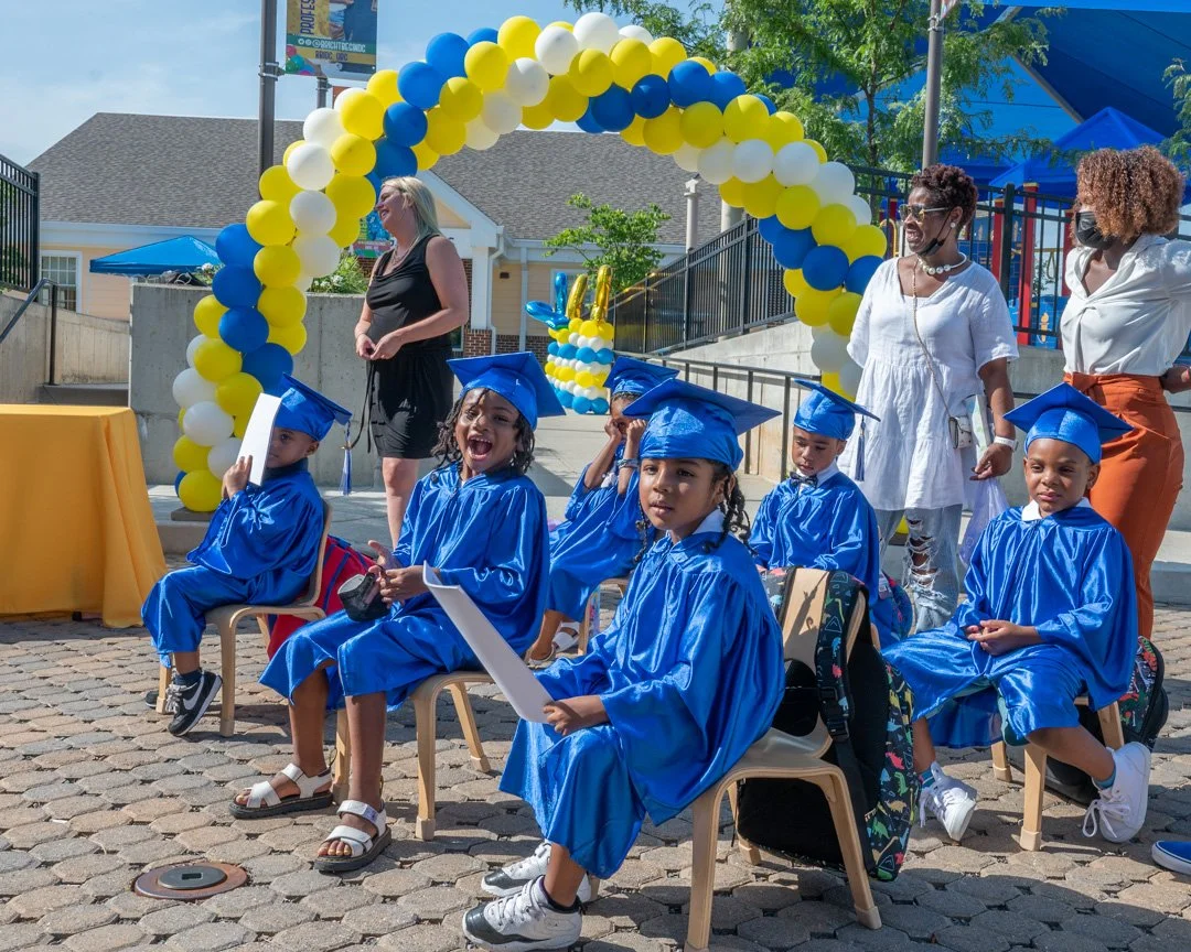 Children wearing blue graduation caps and gowns seated on chairs outdoors during a graduation ceremony, decorated with yellow, blue, and white balloons and a colorful balloon arch.