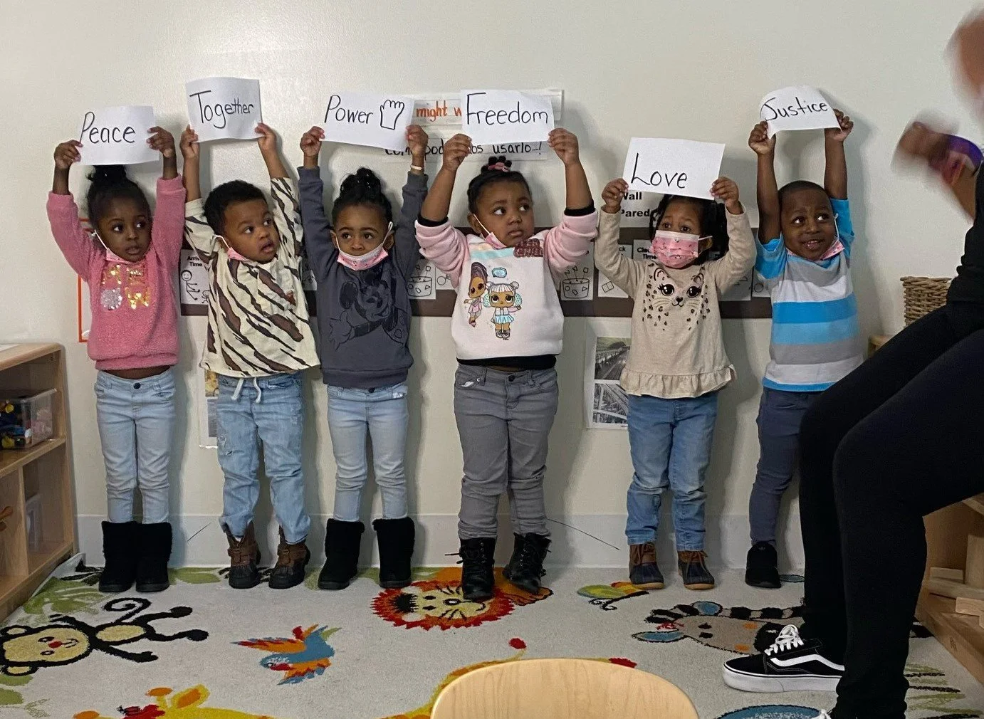 Six young children standing in a classroom holding signs with words like Peace, Together, Power, Freedom, Love, Justice. They are wearing casual clothes and some have masks, with a teacher or adult partially visible on the right side of the image.