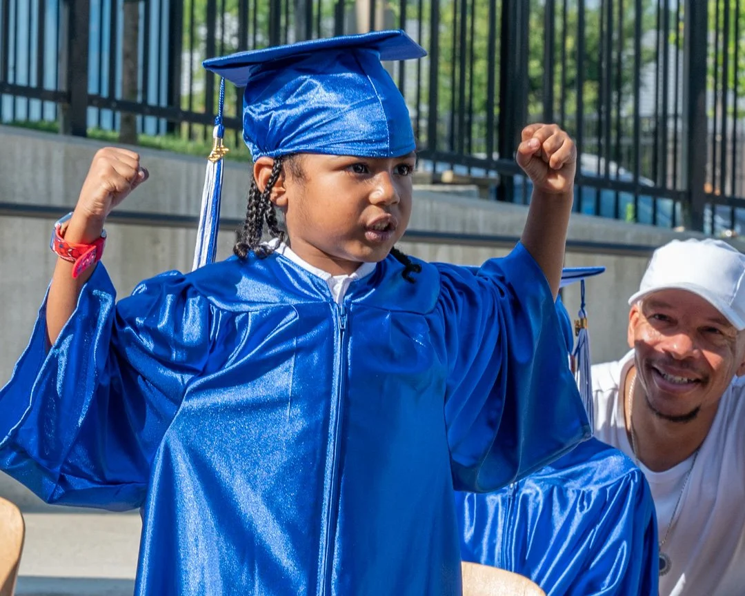 Young girl in blue graduation cap and gown flexing her arms in celebration, standing outside with a man smiling nearby.