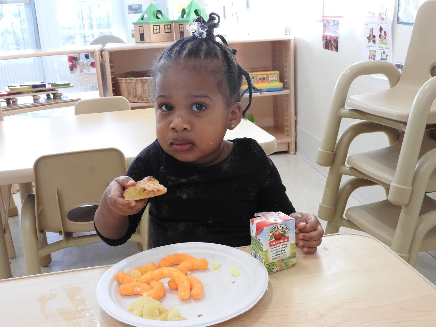 A young girl sitting at a table in a classroom, holding a slice of pizza in her right hand and a carton of apple juice in her left, with a meal of cheese puffs and mashed potatoes on a white paper plate in front of her.