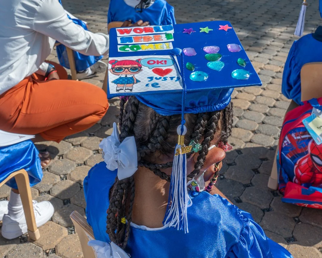 A young girl wearing a blue graduation cap decorated with colorful stickers and drawings, including the words "World We Are Come!" and a cartoon girl with glasses. She has braided hair with white ribbons and is dressed in a blue graduation gown, sitt