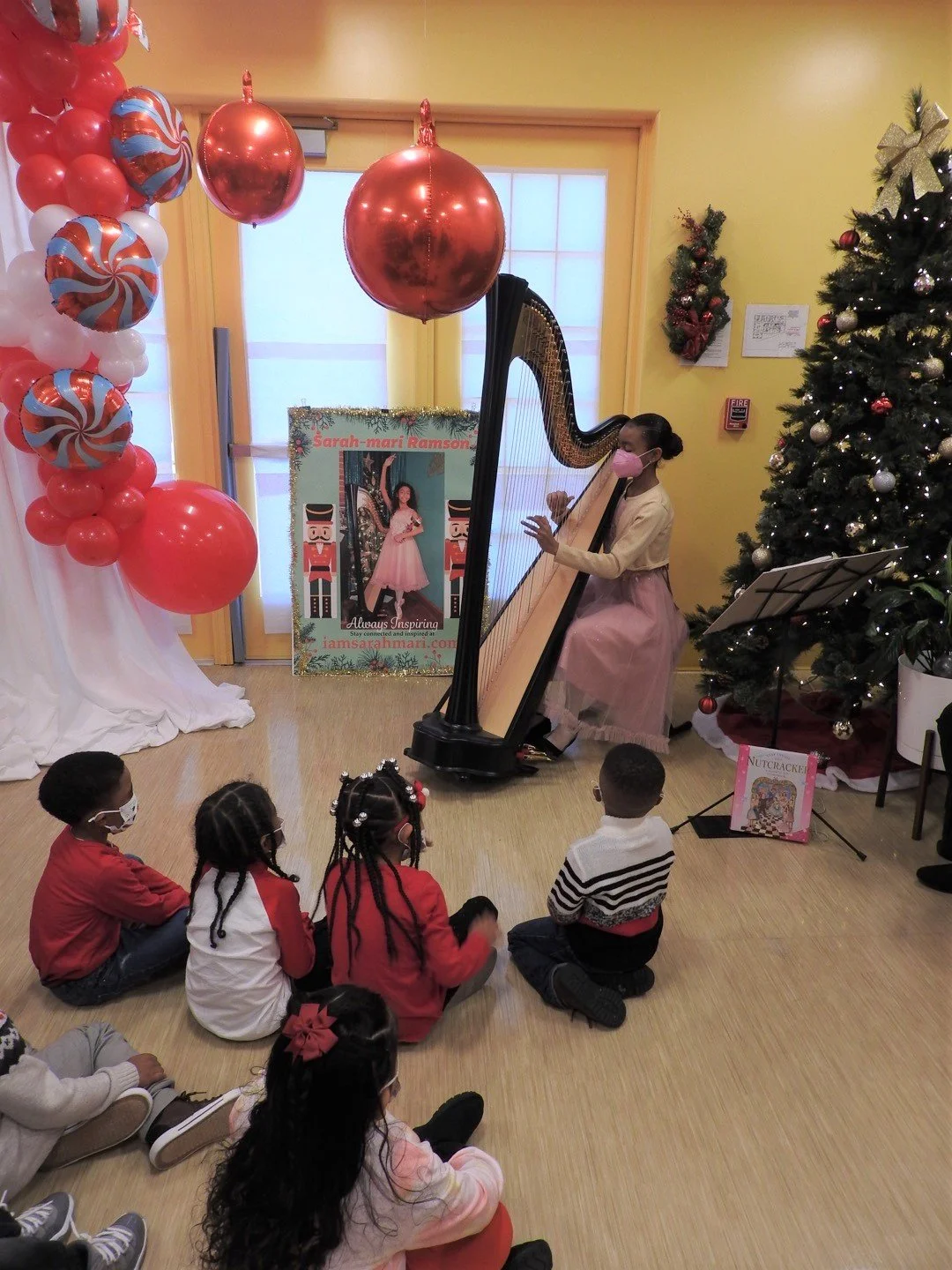 A young girl dressed in a pink tutu and beige jacket plays a harp as children sit on the floor watching her, in a room decorated with Christmas ornaments including balloons, a Christmas tree, and a poster of a girl in a pink dress.