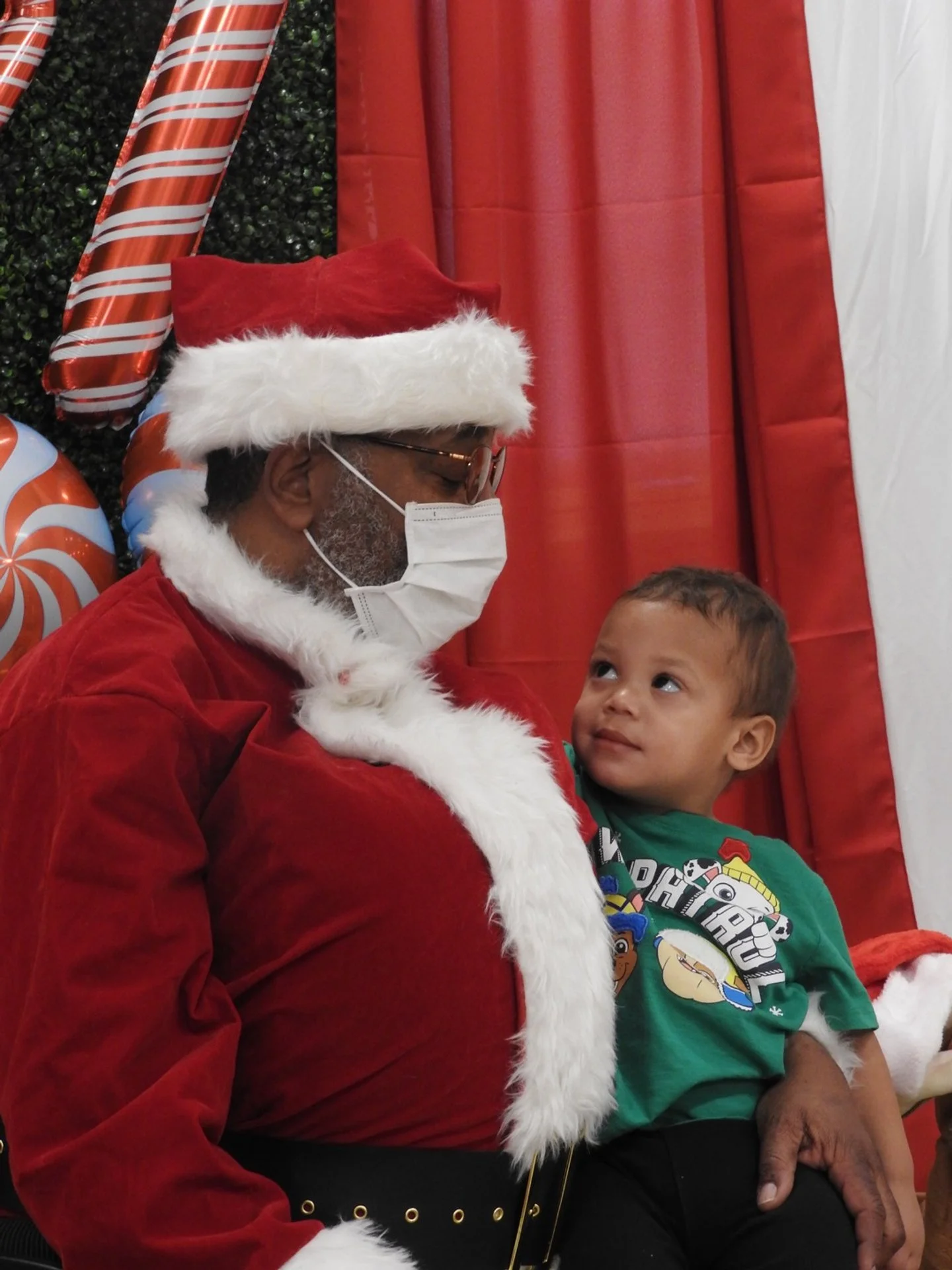 A young boy sitting on a man's lap, looking up at him. The man is dressed as Santa Claus with a red suit, white beard, Santa hat, glasses, and a face mask. The background has red, white, and candy cane decorations.
