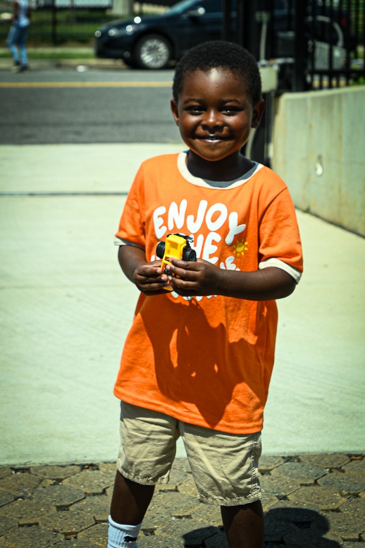 A smiling young boy with dark skin and short curly hair, wearing an orange T-shirt with white text that says 'Enjoy the', khaki shorts, and striped socks, holding a yellow toy truck outdoors on a sunny day.