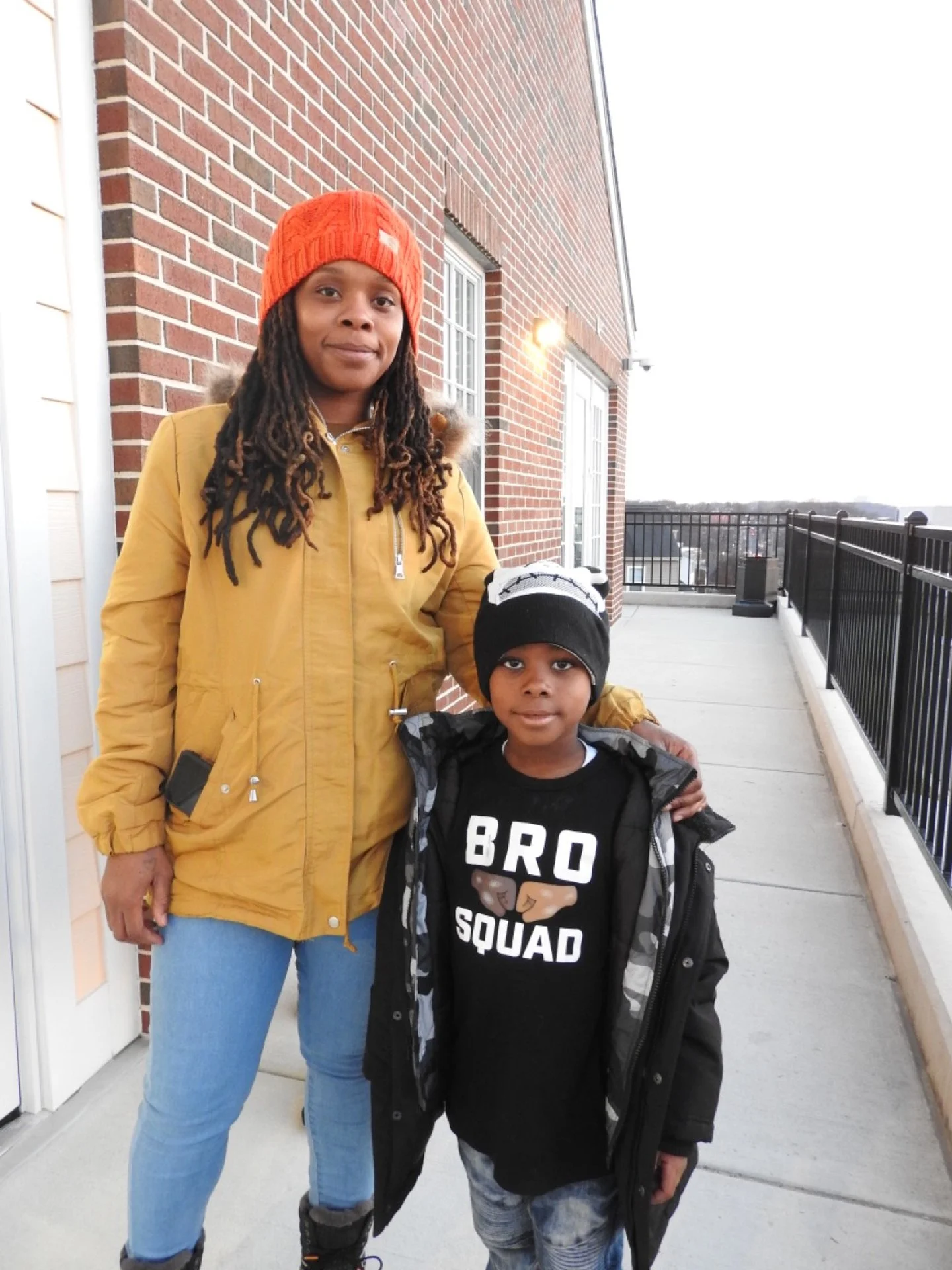 A woman and a young boy standing on a balcony outside a brick building. The woman is wearing a yellow jacket, light blue jeans, and an orange beanie. The boy is wearing a black beanie, a black T-shirt that says 'BRO SQUAD' with a graphic, and a black