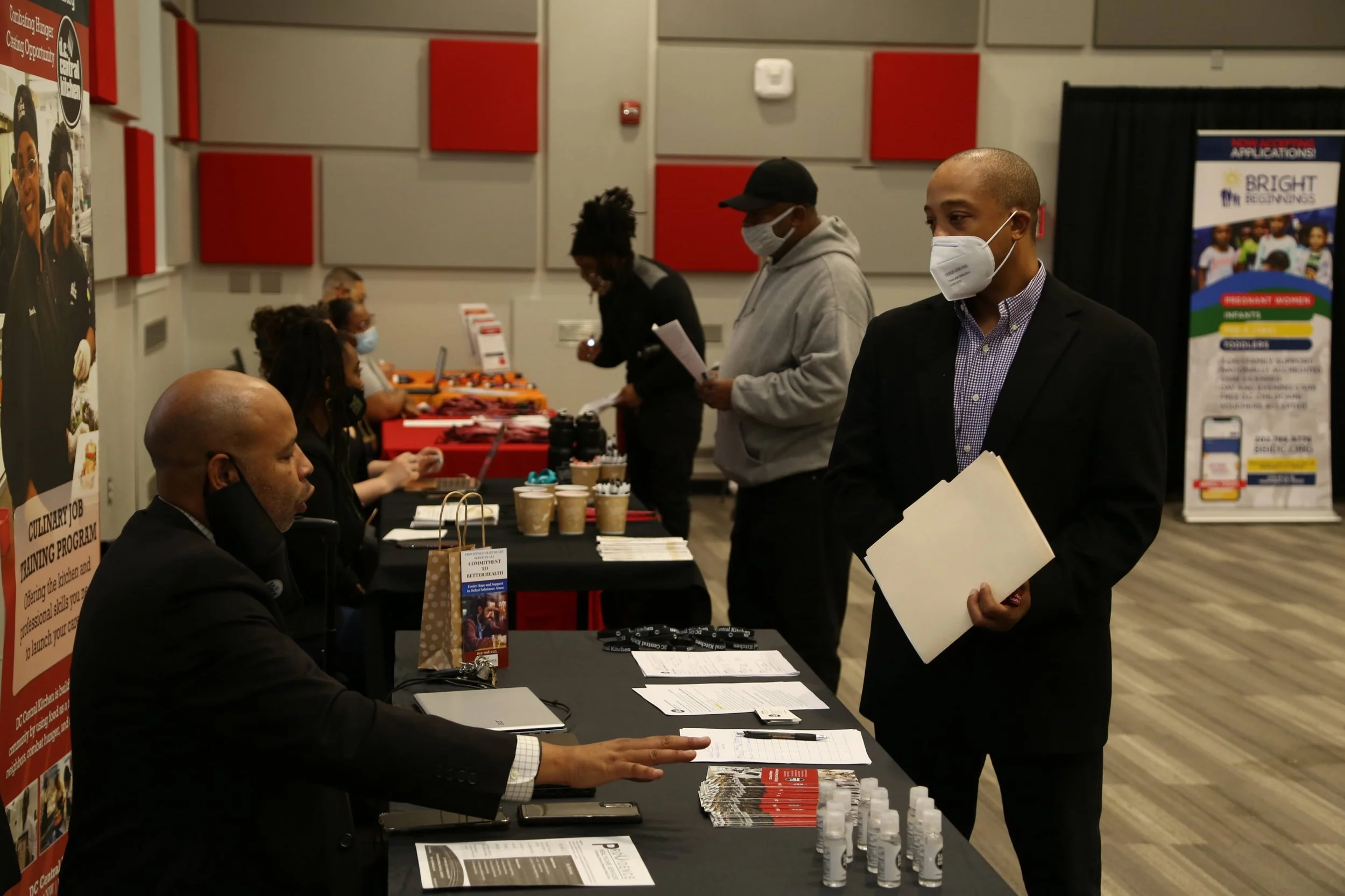 People attending a career fair or networking event, wearing masks, and engaging at a table with informational materials, hand sanitizer bottles, and promotional items in a room with red and gray wall panels.