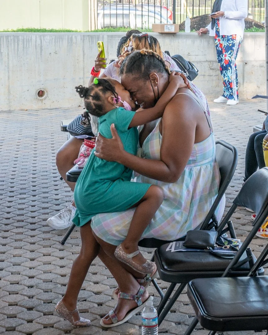 A woman and a young girl are sharing an embrace, touching foreheads and smiling, while sitting on chairs in an outdoor setting. Several other people are present in the background, including a woman standing and using a phone. The atmosphere appears j
