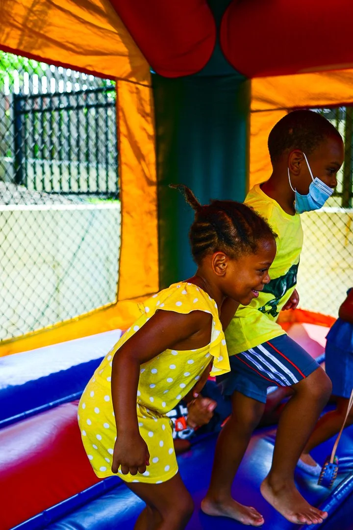 Children playing inside a colorful bounce house, with yellow and orange walls, one girl in a yellow polka dot dress and a boy in a yellow shirt and blue shorts, both smiling and jumping.