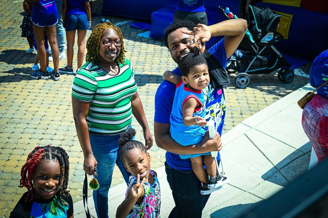 A smiling group of six people, including children and adults, gathered outdoors on a sunny day. The group includes a woman with glasses, a man holding a young boy, and three girls, all dressed casually. The background shows other people, a stroller, 