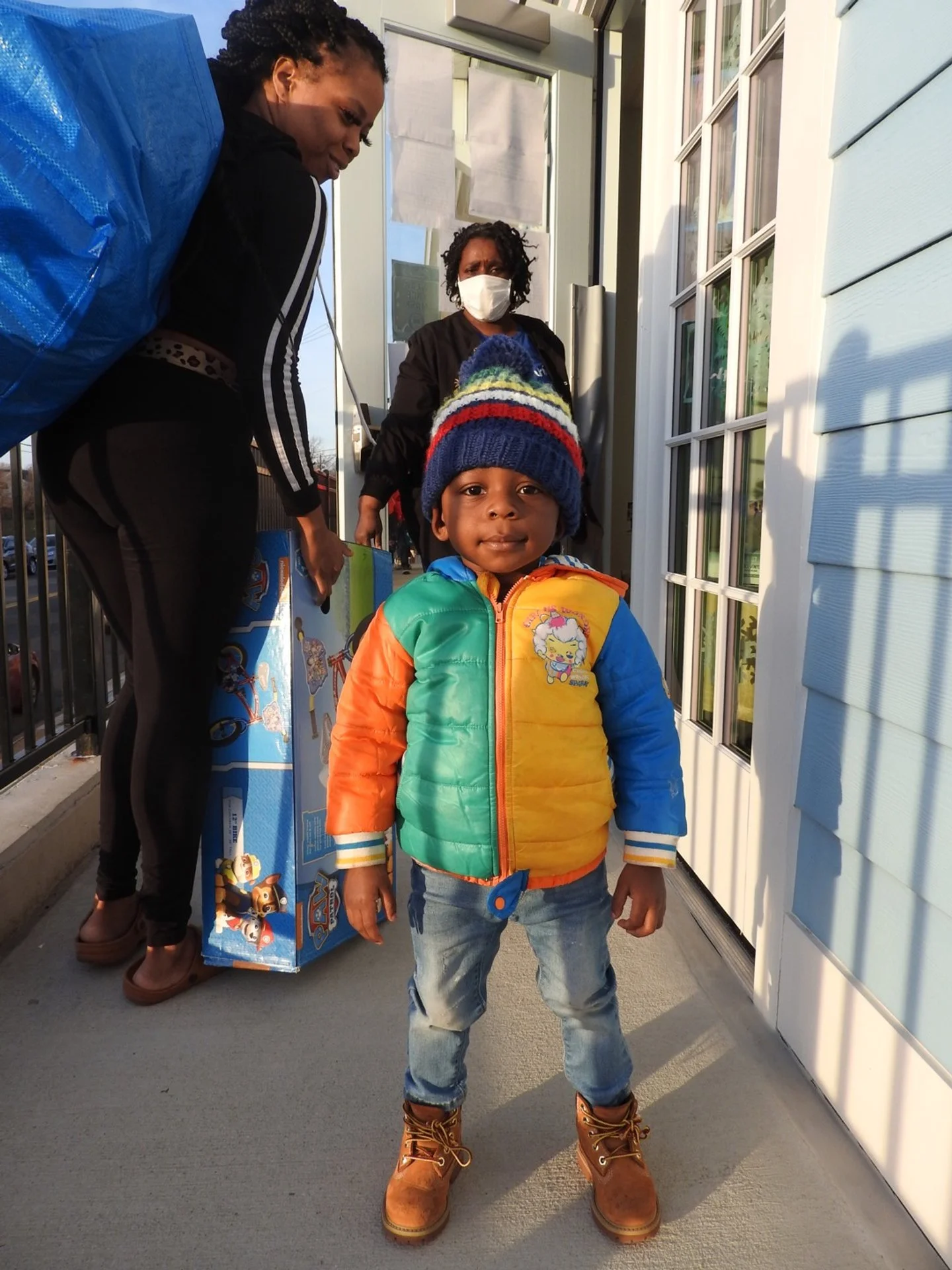 Young boy wearing a colorful jacket, jeans, and a knit hat standing outdoors in front of a house, with two women in the background, one with a mask, and a large box with characters on it.