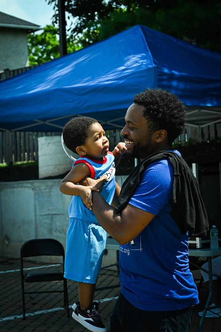 A man holding a young boy up in the air outside under a blue canopy. The boy appears to be upset or crying, while the man is smiling.