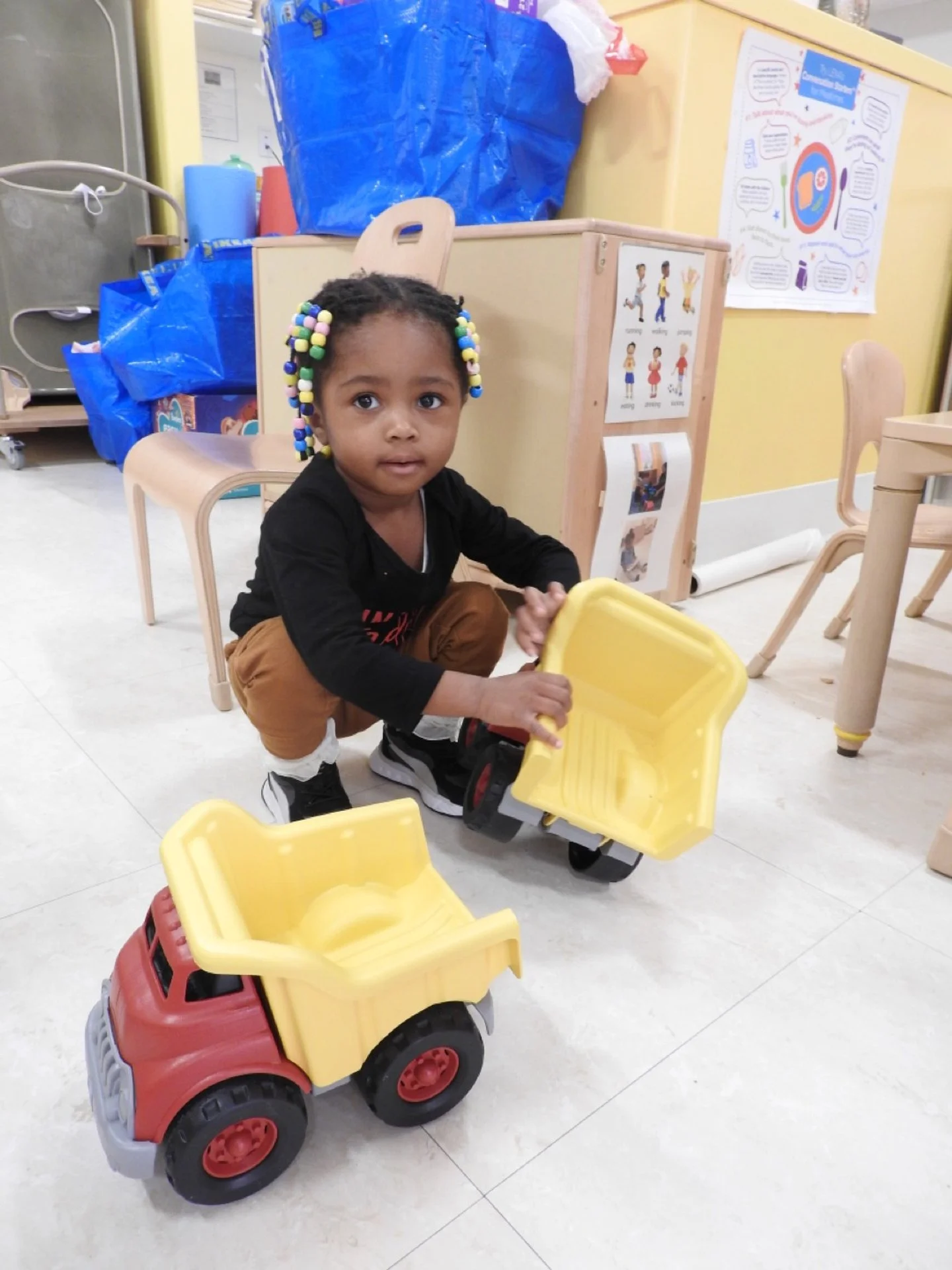 A young girl with colorful beaded hair is playing with a yellow and red toy dump truck in a classroom setting.