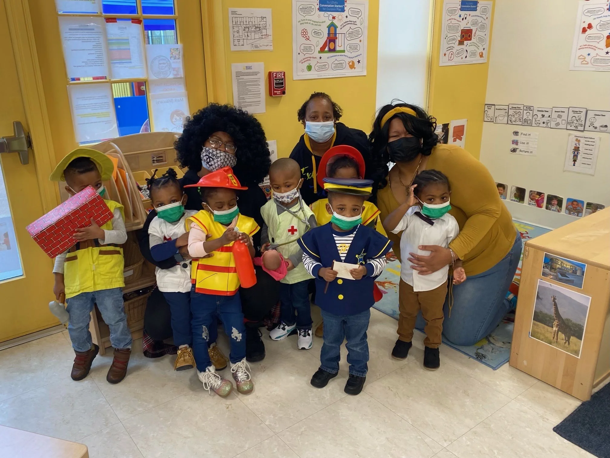 A group of young children dressed in costumes and wearing face masks, posing with three adults in a classroom. The children appear to be dressed as various professionals or characters, and are holding props. The classroom has colorful posters on the 