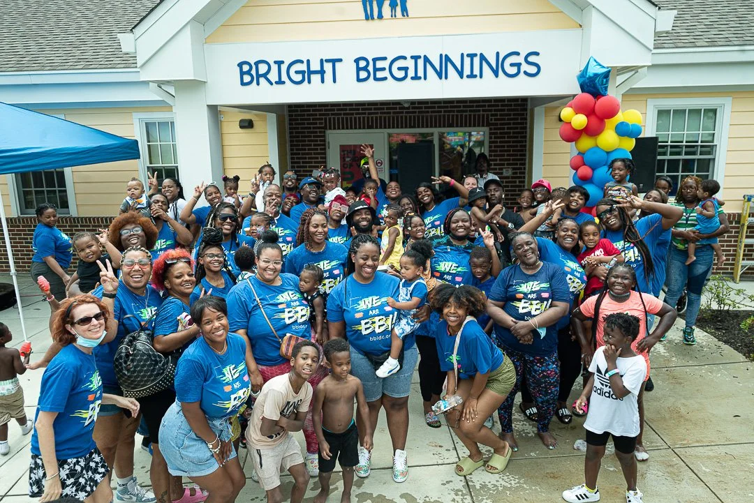Group of children and adults smiling in front of a building with a sign that reads 'Bright Beginnings,' decorated with balloons, during a community event.
