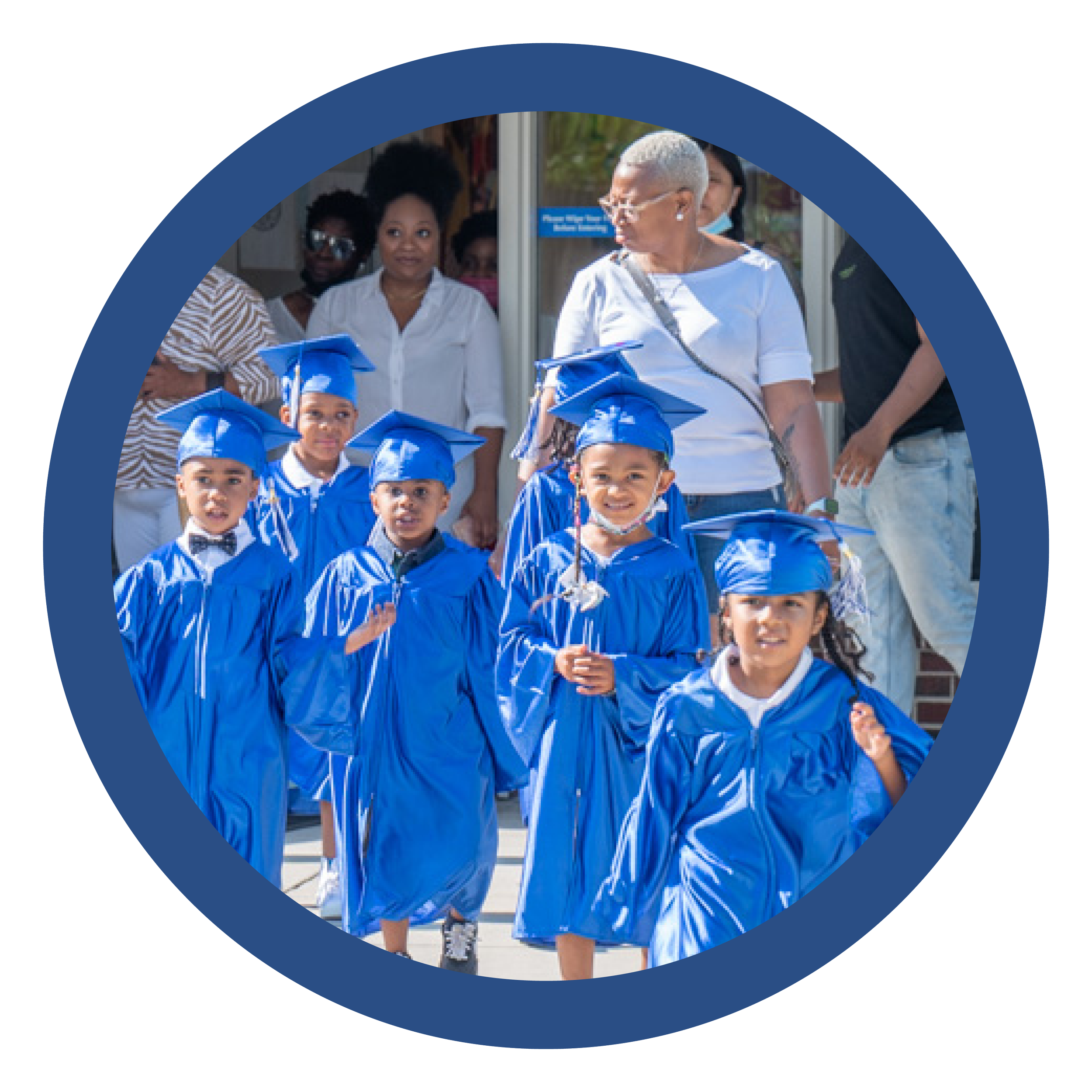 Group of young children in blue graduation gowns and caps walking outside, with adults watching in the background.